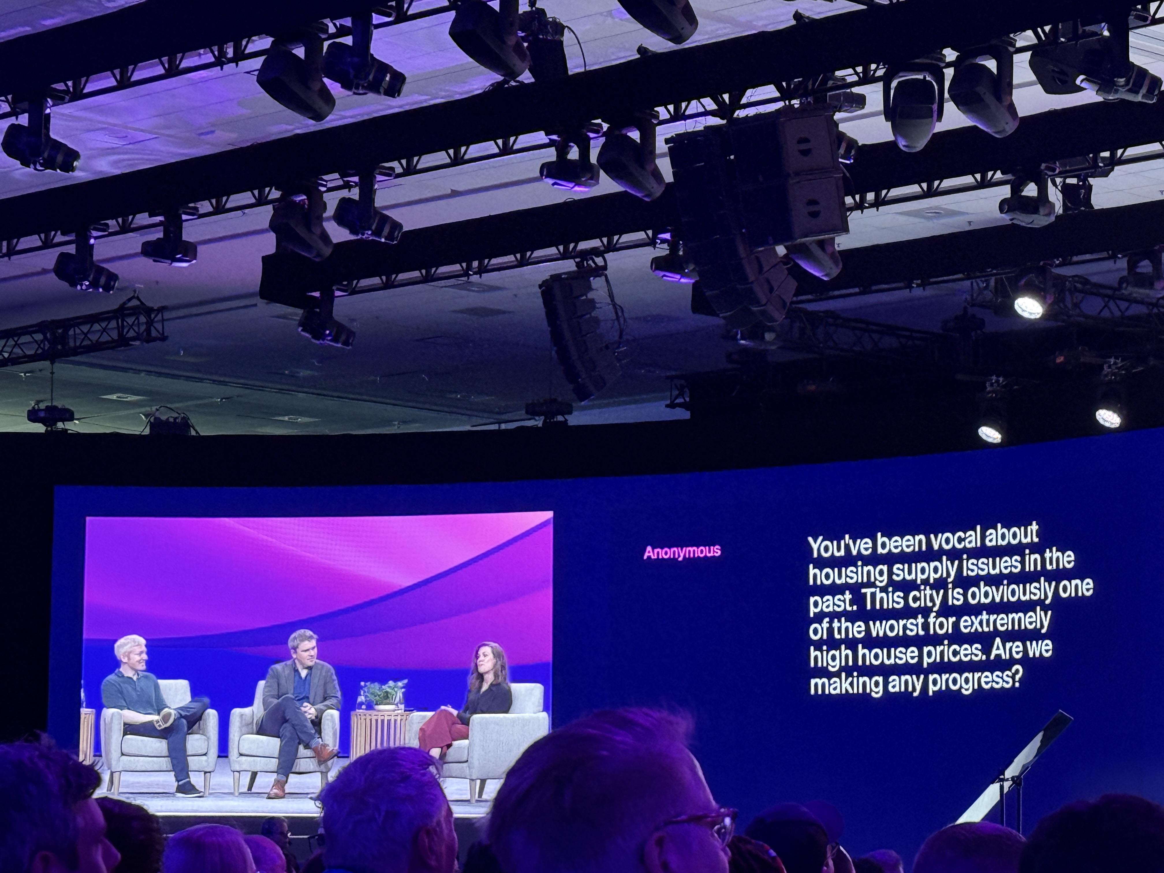 Fireside chat at Stripe Sessions 2025 with three speakers seated on stage in front of a purple backdrop, alongside a large screen displaying an audience question about housing supply and affordability in San Francisco.