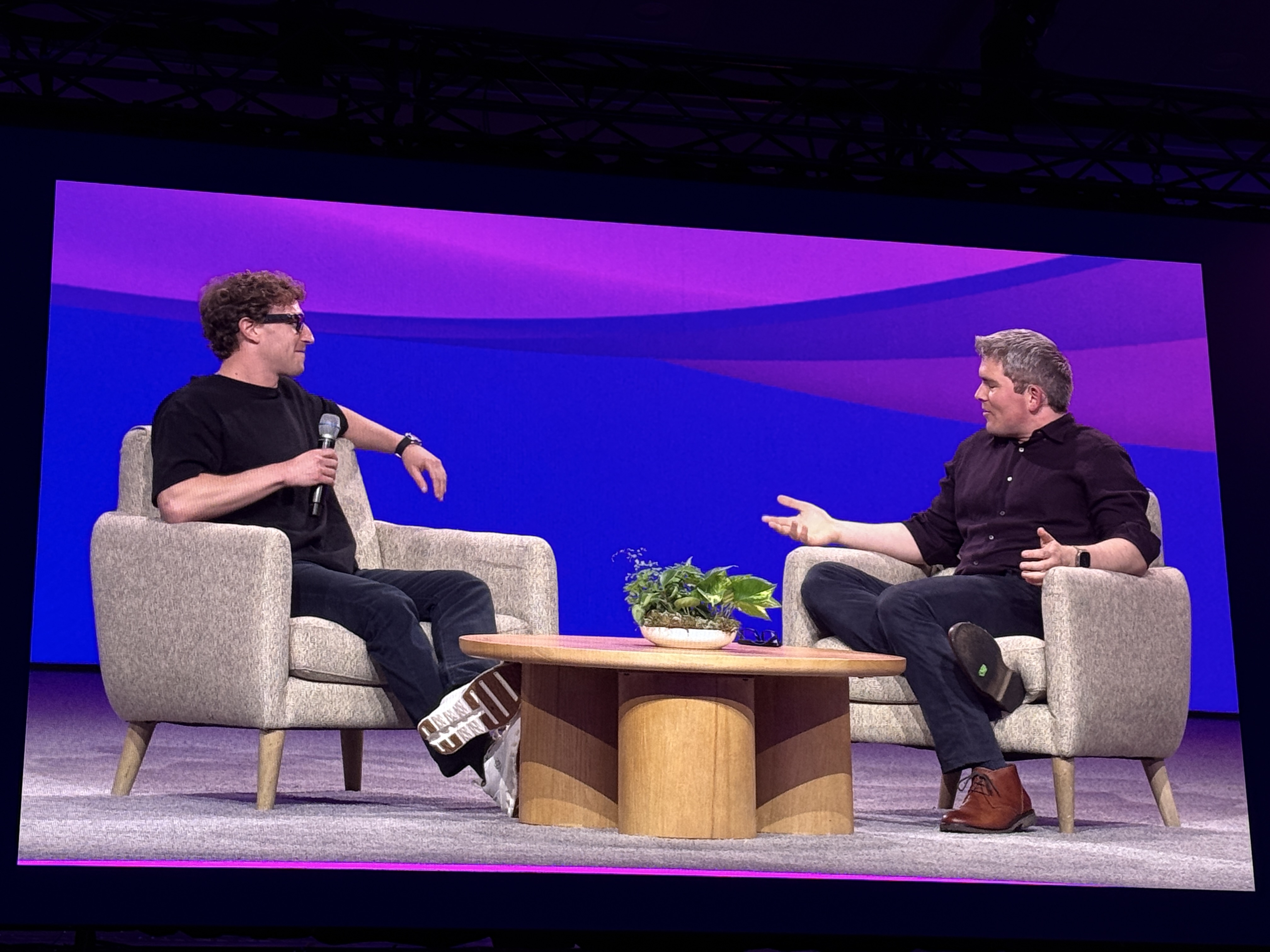 Fireside chat at Stripe Sessions 2025 with two speakers seated in armchairs on stage, one holding a microphone. They are in conversation against a purple backdrop, with a small wooden table and plant between them.