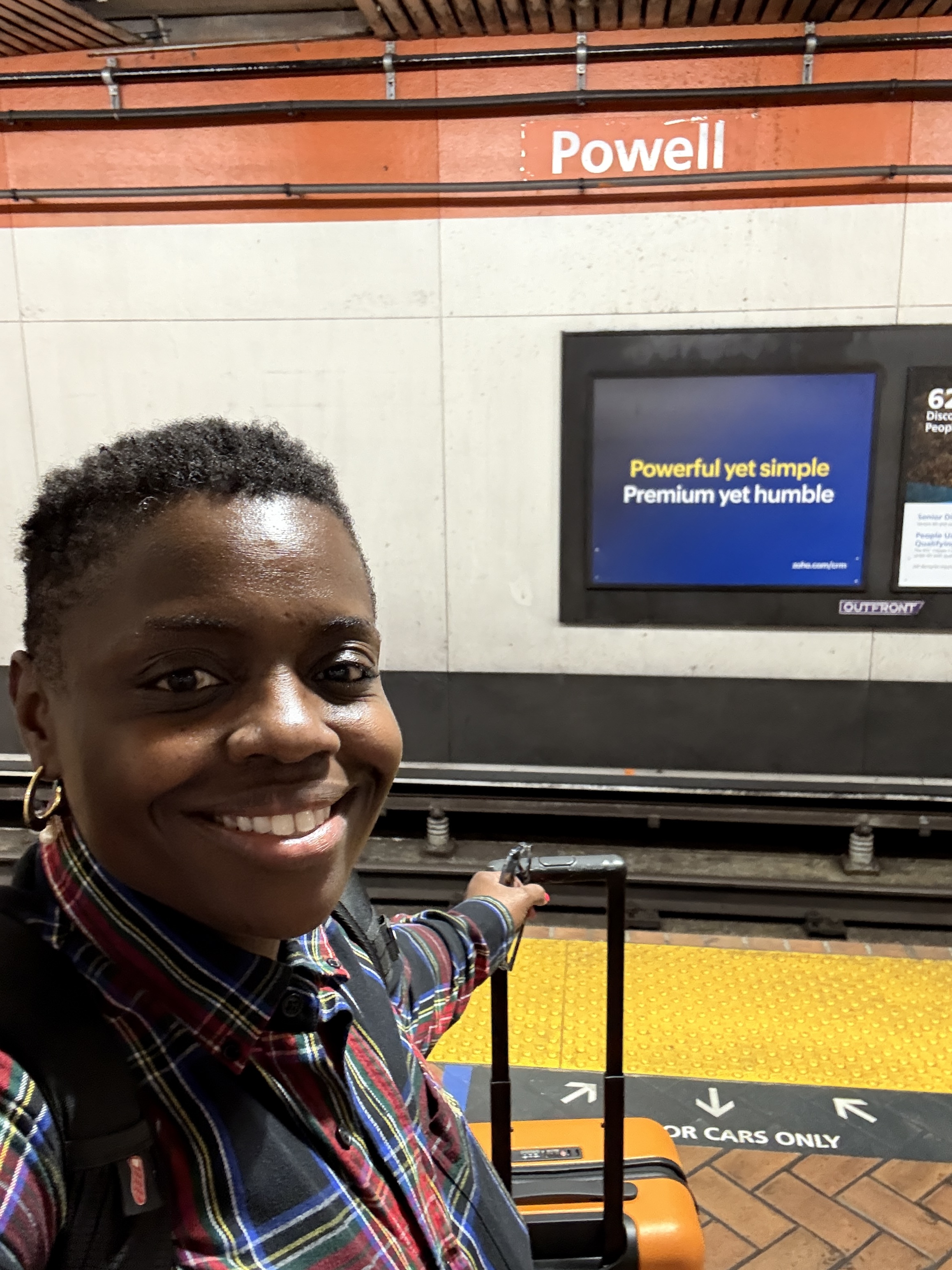 Person smiling with suitcase on the Powell Street BART station platform in San Francisco, pointing toward the tracks. Behind them is a digital ad with the words ‘Powerful yet simple. Premium yet humble.