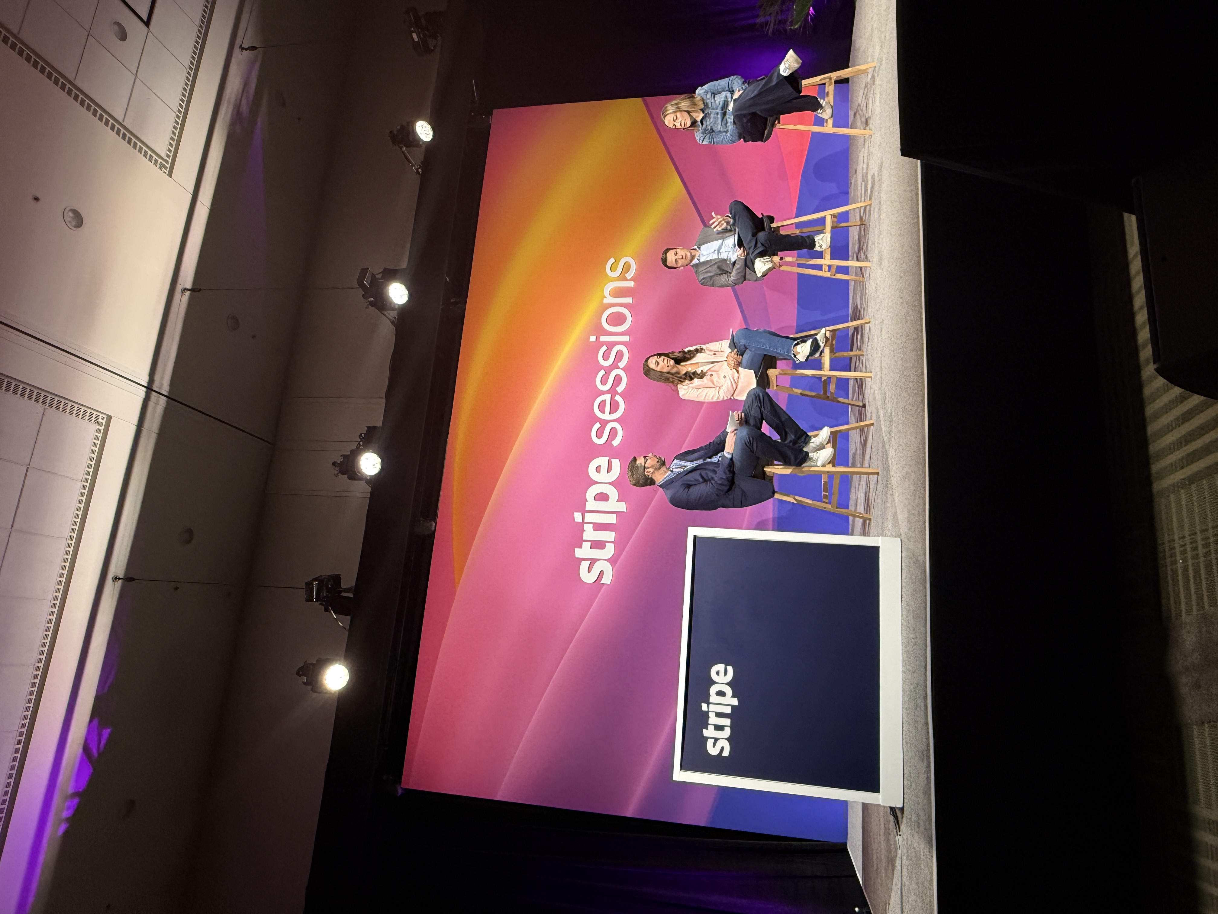 Panel discussion at Stripe Sessions 2025 with four speakers seated on stage in front of a vibrant pink and orange backdrop reading ‘stripe sessions,’ alongside a podium with the Stripe logo.