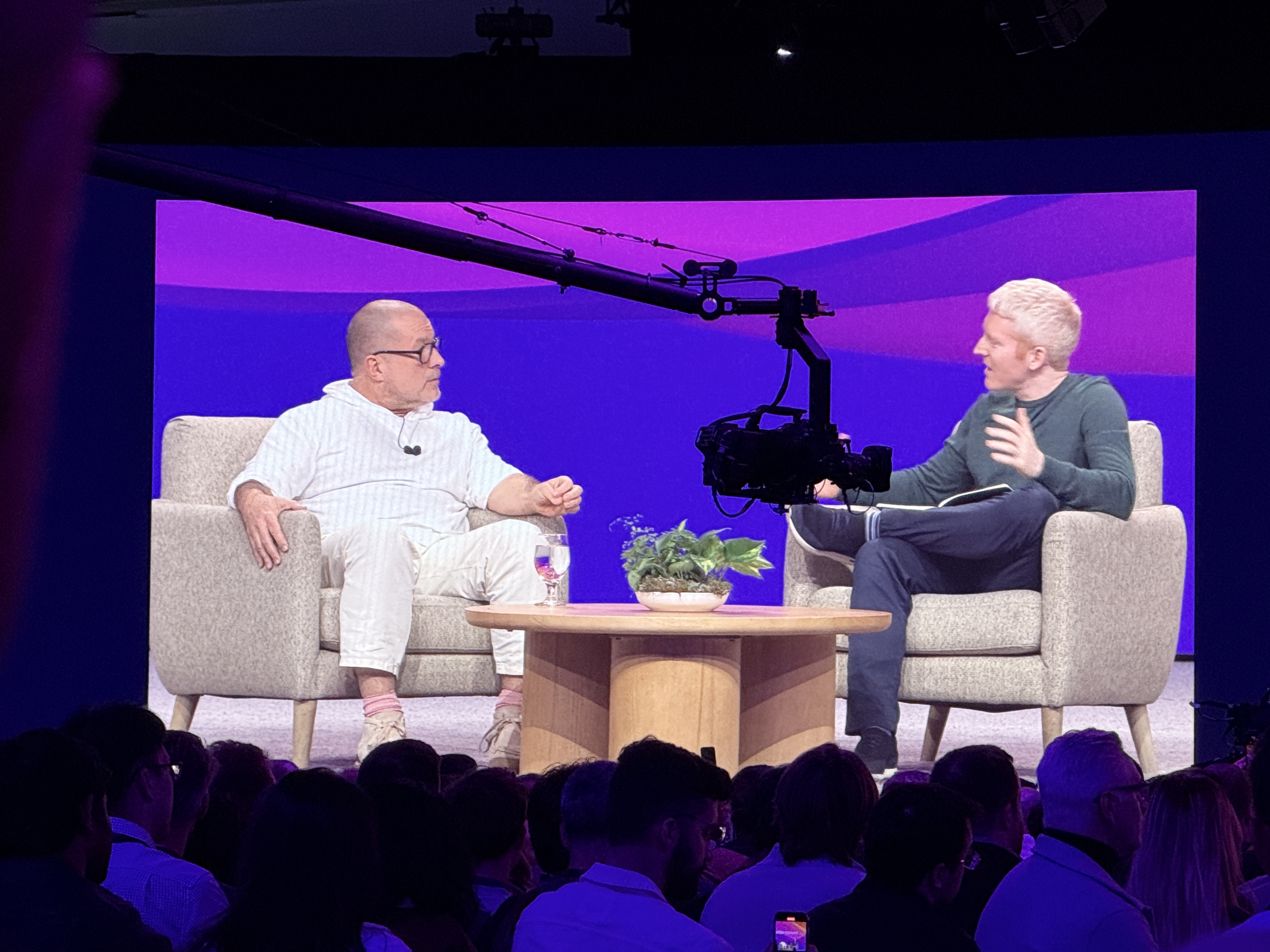 Fireside chat at Stripe Sessions 2025 with two speakers seated in armchairs on stage, one wearing a white outfit and the other in a dark green shirt, in conversation against a purple backdrop with a camera rig visible in front of them.