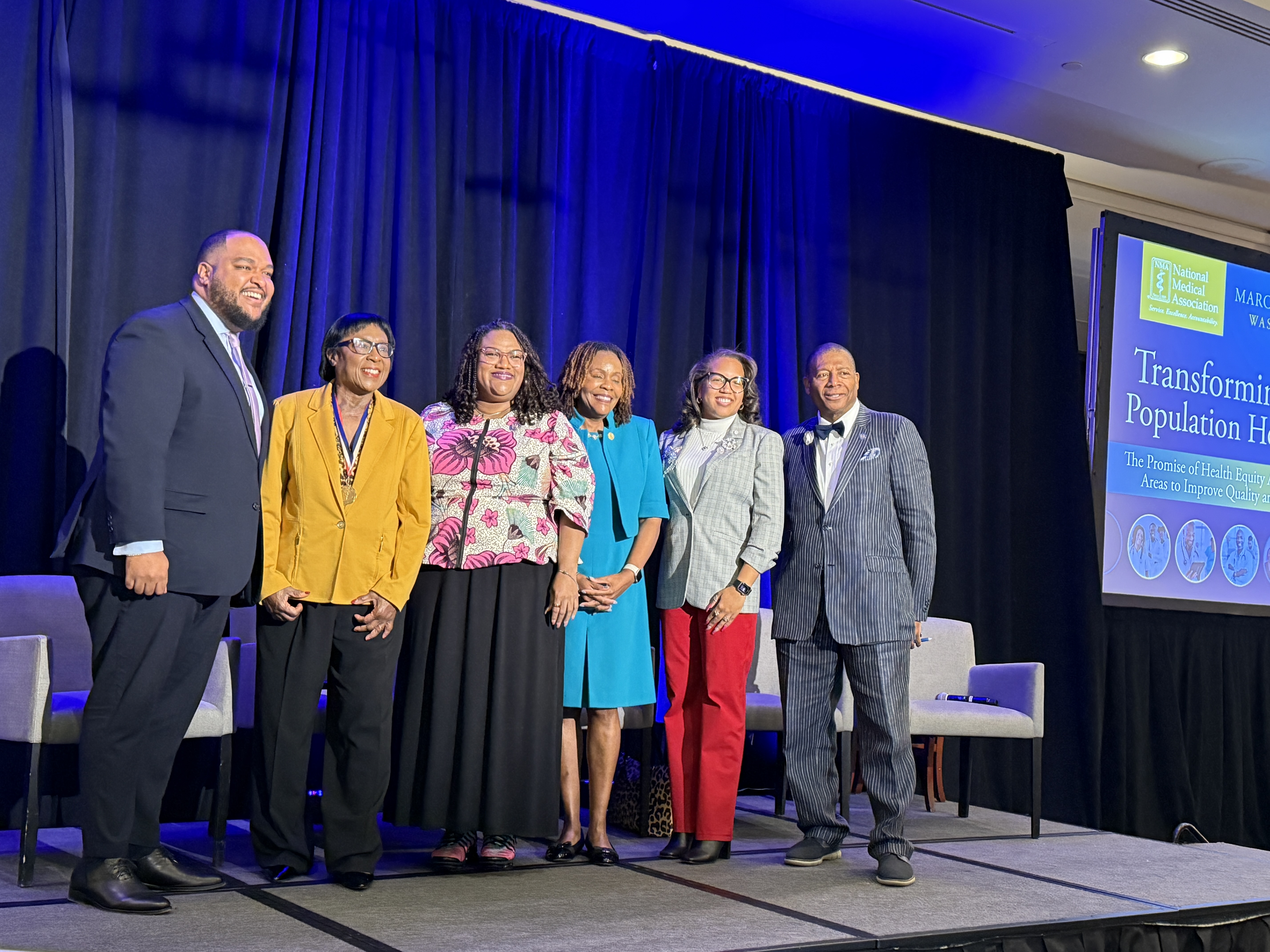 Group of six speakers on stage at the NMA Colloquium, smiling together in front of a backdrop that reads ‘Transforming Population Health: The Promise of Health Equity in Key Areas to Improve Quality of Care.