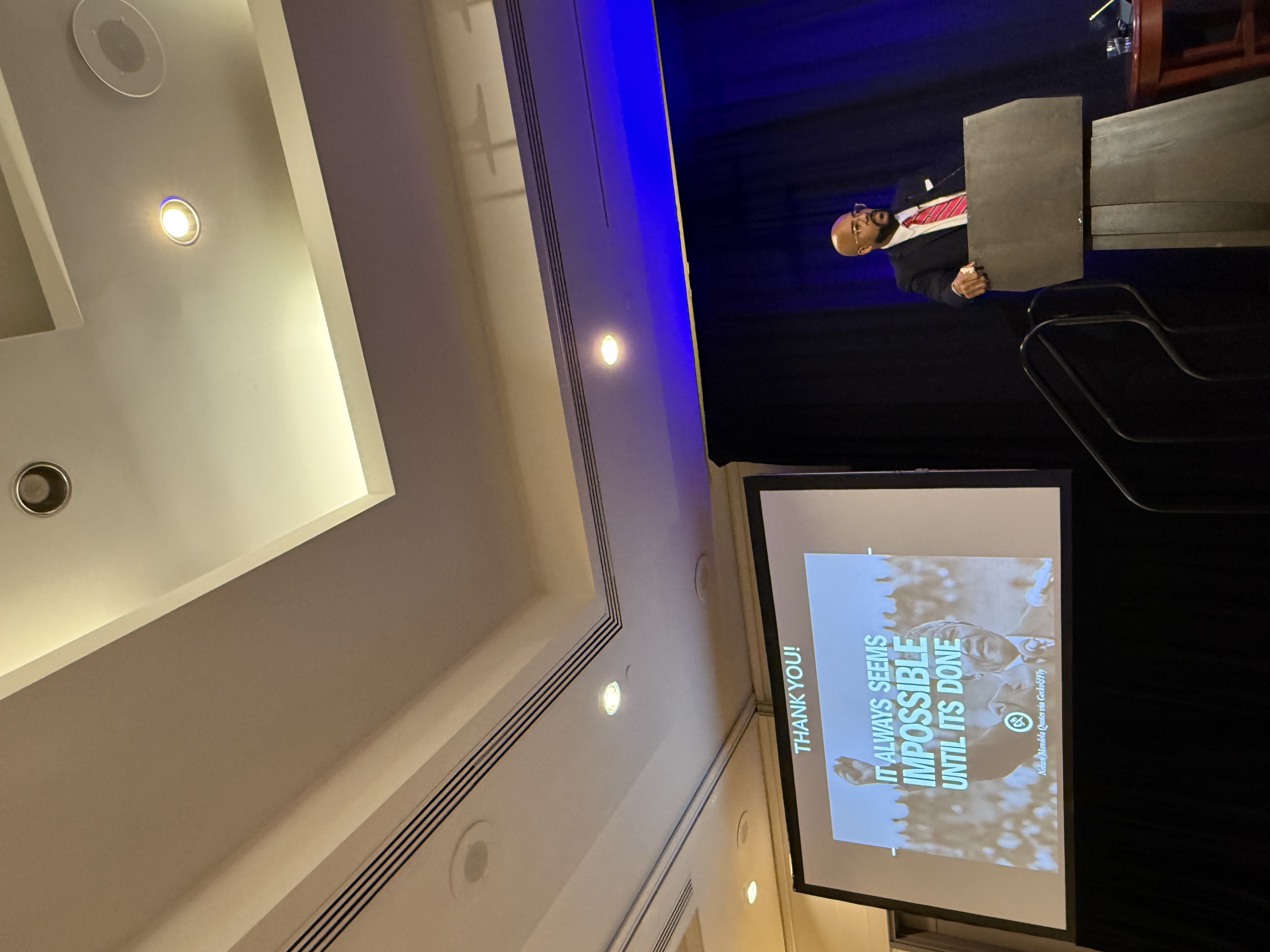 Speaker at the NMA Colloquium stands at a podium beside a screen projecting a Nelson Mandela quote that reads, ‘It always seems impossible until it’s done.’