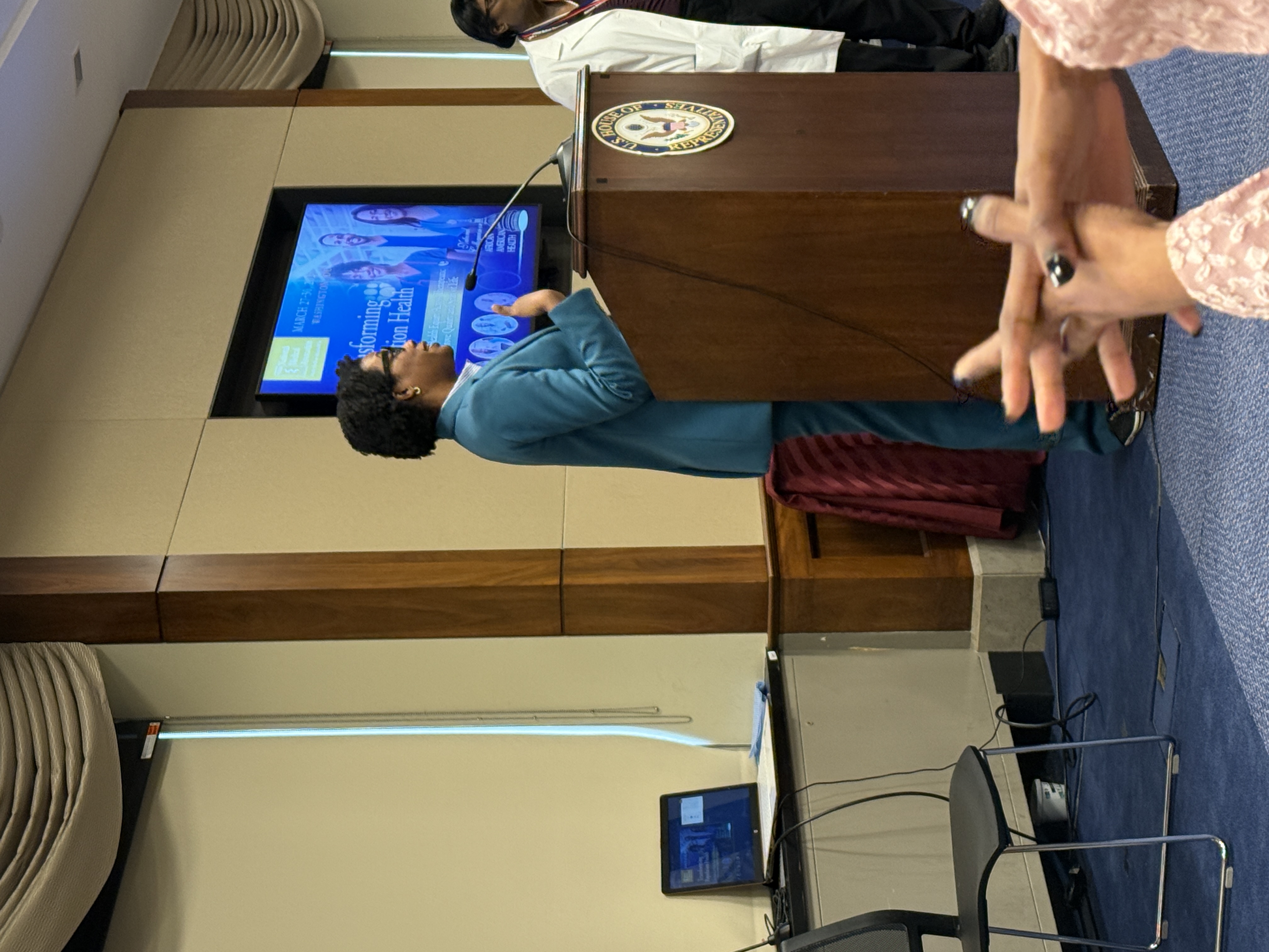 Representative Lauren Underwood of Illinois speaks at the NMA Colloquium from a podium with the U.S. House of Representatives seal, during a session on transforming population health, with a digital event banner visible in the background.