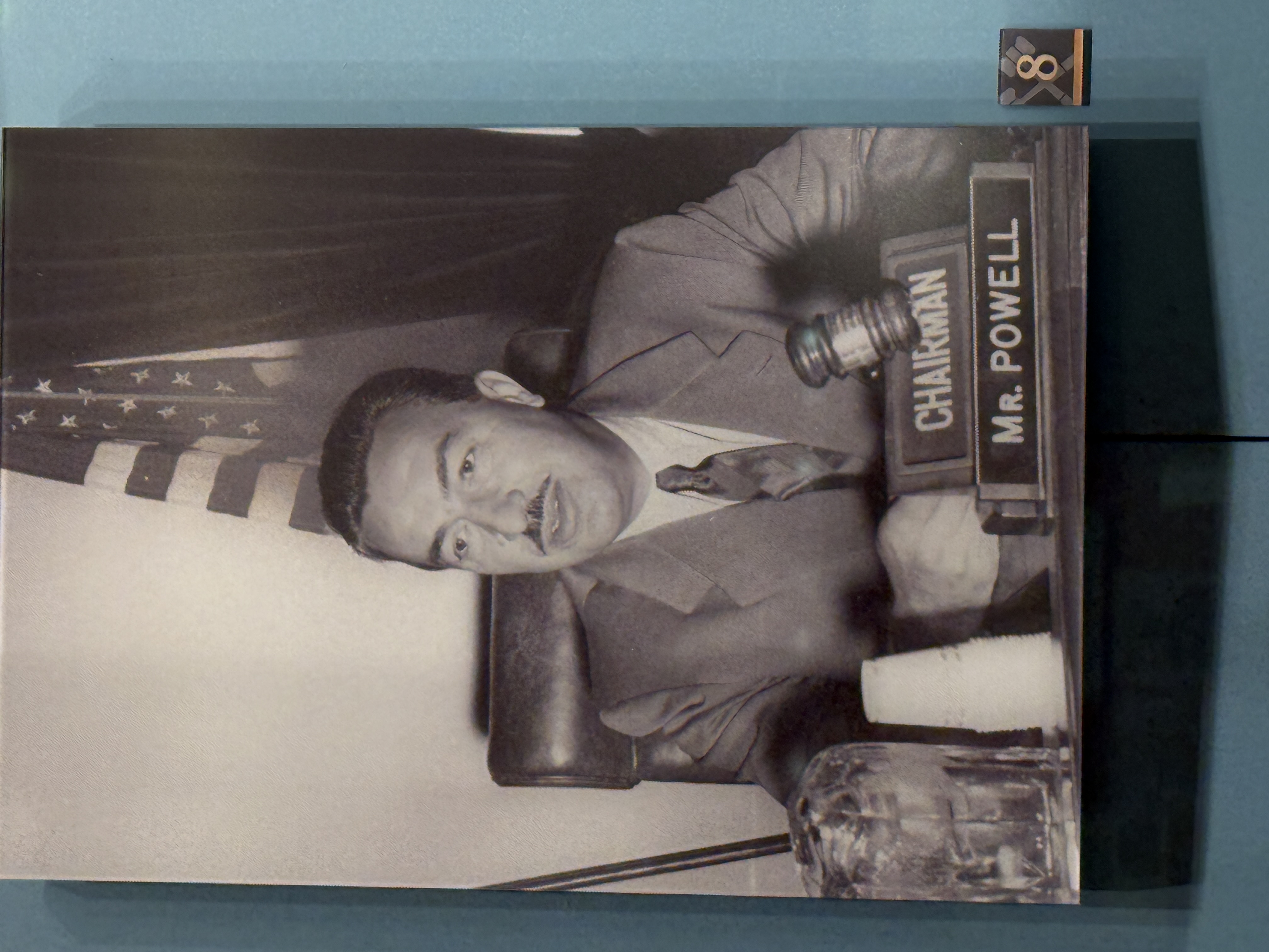 Black-and-white photograph of Adam Clayton Powell, Jr. seated at a desk with a gavel, nameplate reading ‘Chairman Mr. Powell,’ and an American flag in the background, displayed as part of a Capitol exhibit.
