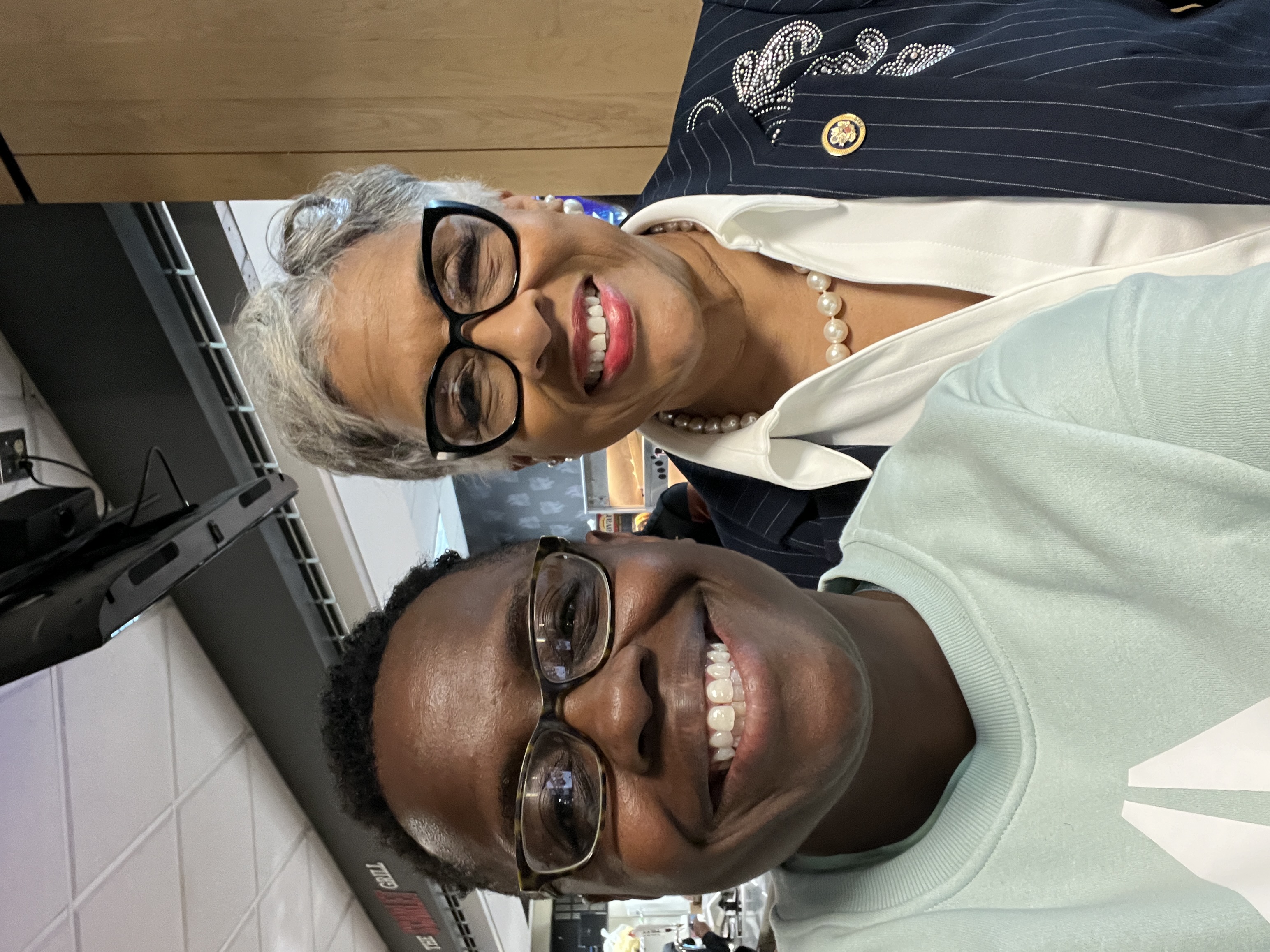Two people smile for a close-up selfie indoors. The person on the left, a seasoned woman of executive and congressional leadership with short gray hair and black glasses, wears a navy pinstripe blazer with decorative embellishments, a white collared shirt, and a pearl necklace. The person on the right, a younger individual, a constituent within her district, with short hair and glasses, wears a light-colored sweatshirt. Both appear happy, standing close together in a well-lit space with other people and signage visible in the background.