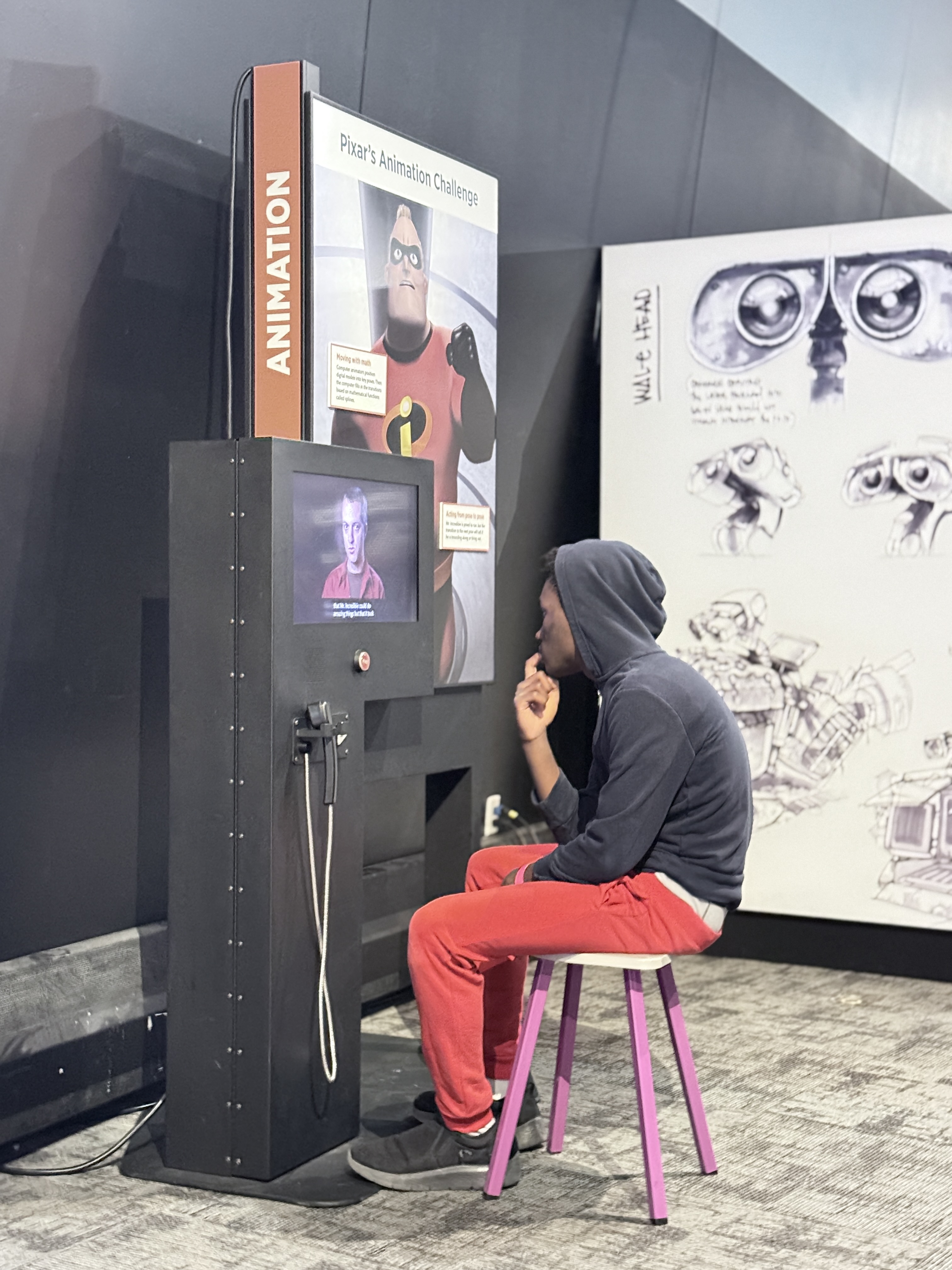A boy sits on a small stool at an interactive Pixar animation exhibit, watching a video screen while resting his chin on his hand in a thoughtful pose. The display features Mr. Incredible from The Incredibles and signage reading “Pixar’s Animation Challenge,” with sketches of animated robot characters on the wall behind him in a science and creativity museum setting. A boy sits on a small stool at an interactive Pixar animation exhibit, watching a video screen while resting his chin on his hand in a thoughtful pose. The display features Mr. Incredible from The Incredibles and signage reading “Pixar’s Animation Challenge,” with sketches of animated robot characters on the wall behind him in a science and creativity museum setting.