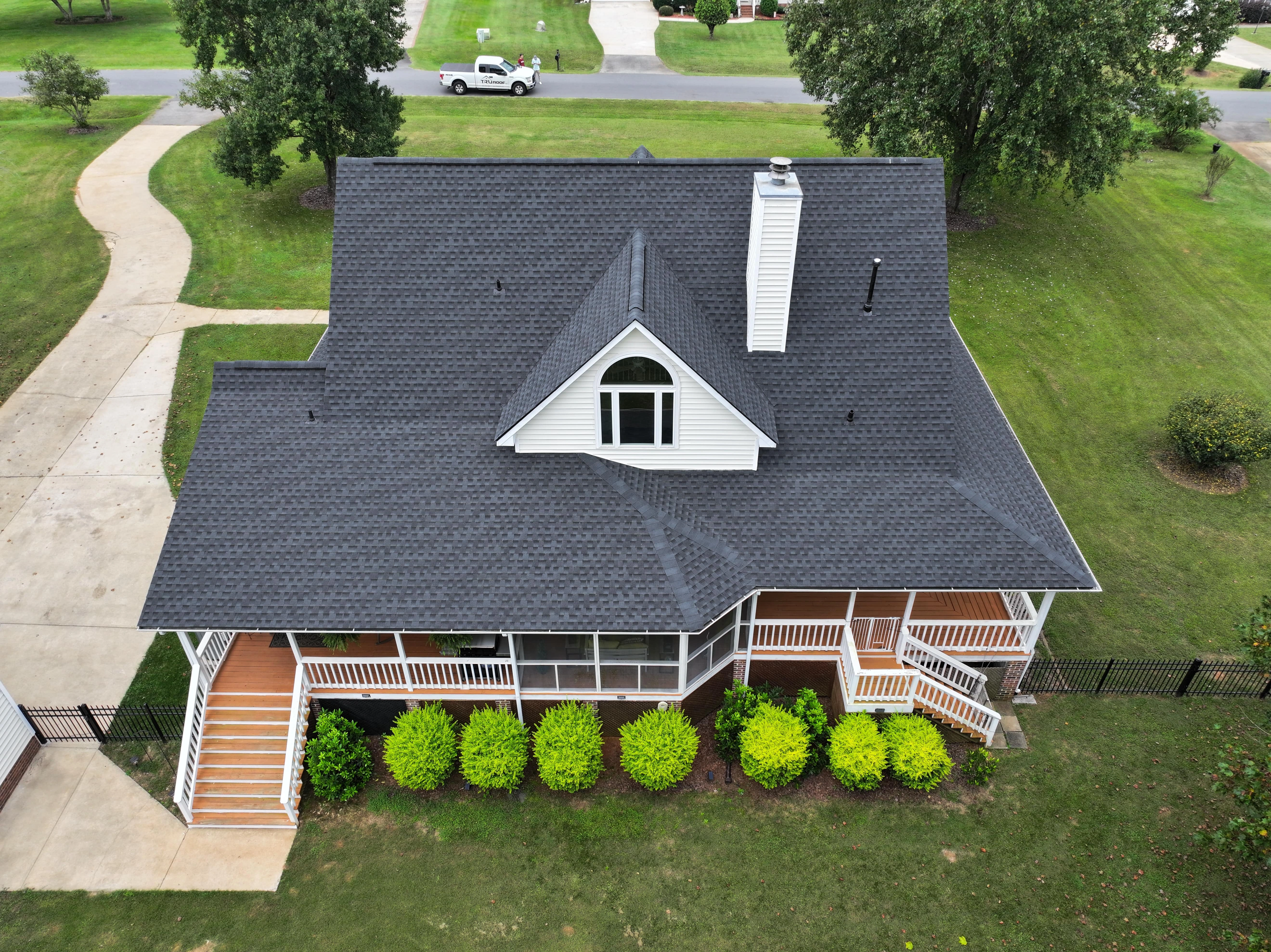 Aerial view of a two-story house with a black shingle roof, white exterior, wraparound porch, and surrounding green lawn and shrubs.
