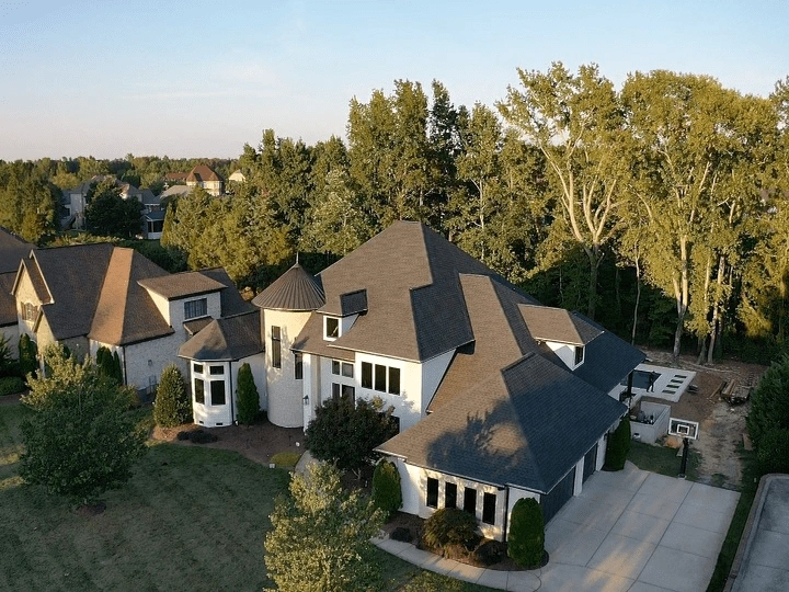 Aerial view of a modern house with a well-maintained shingle roofing system featuring multiple gabled sections, surrounded by a lush green yard, trees, and a concrete driveway.