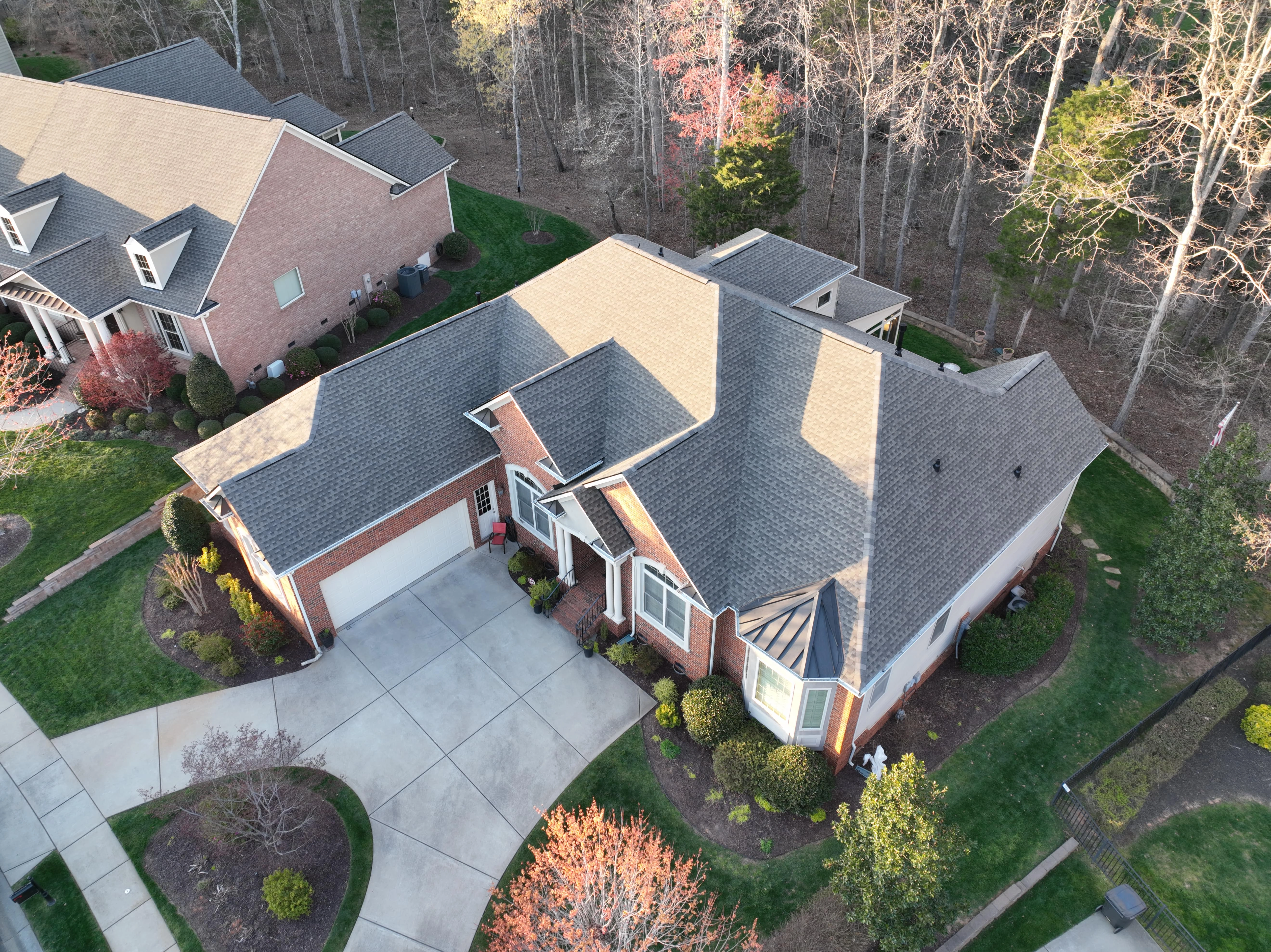 Aerial view of a brick house with a well-maintained shingle roofing featuring multiple gabled sections, surrounded by a landscaped yard with trees and a concrete driveway.