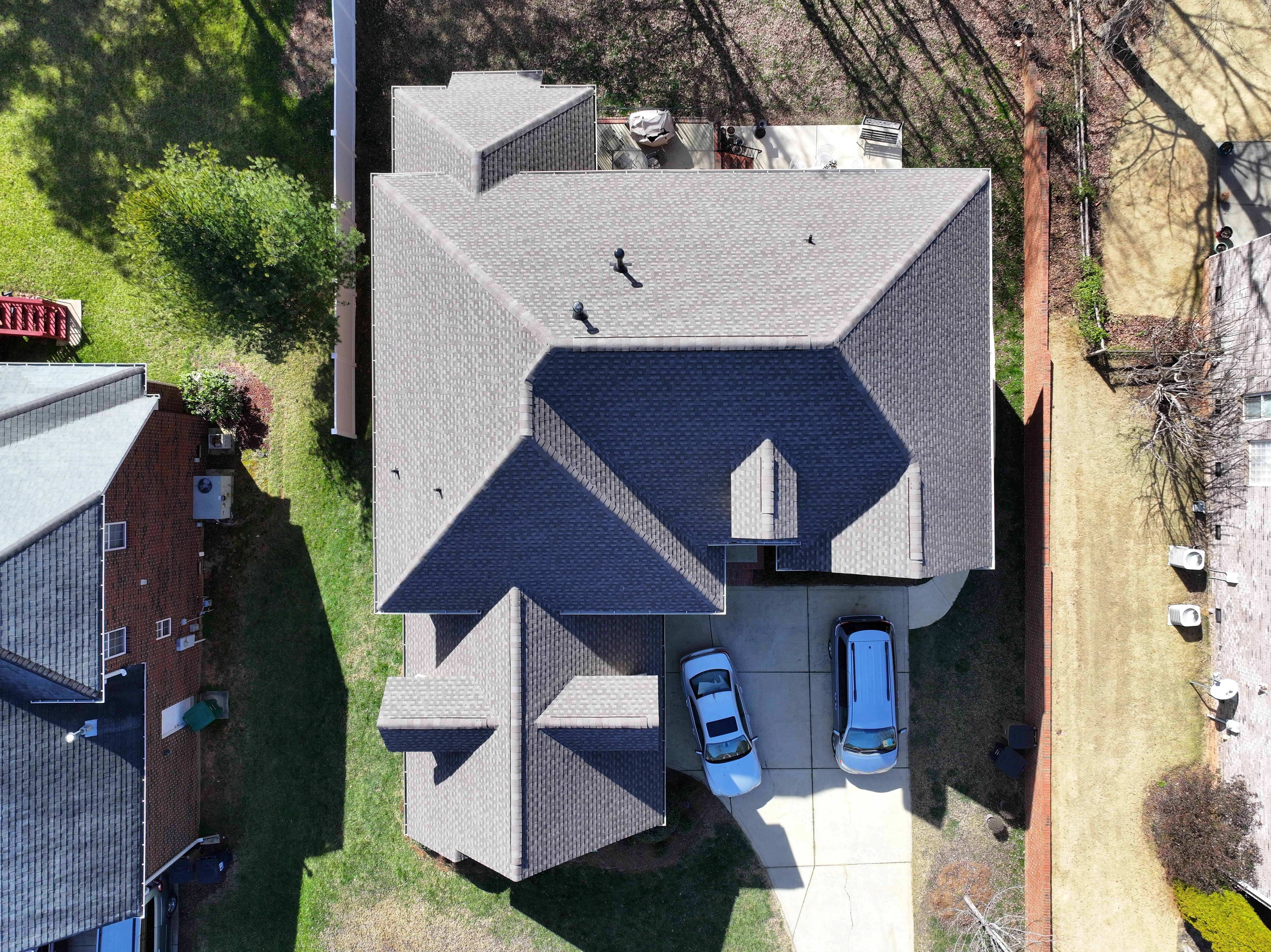 Aerial view of a suburban house with a shingle roofing, driveway, and two parked cars. Neighboring homes and yards are visible on either side.