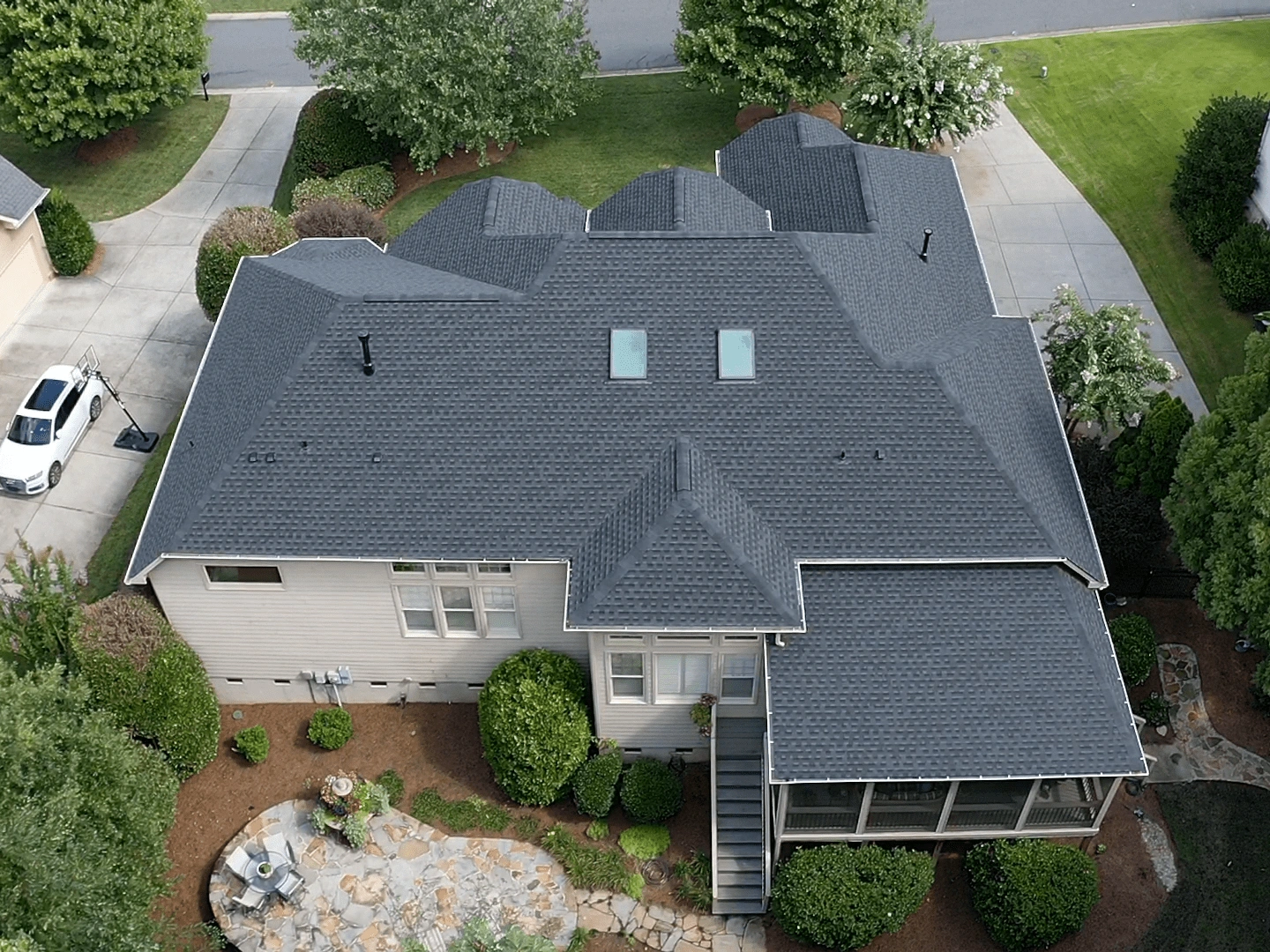 Aerial view of a house with a dark shingle roofing system featuring multiple gabled sections and skylights, surrounded by a landscaped yard with a driveway and patio area.