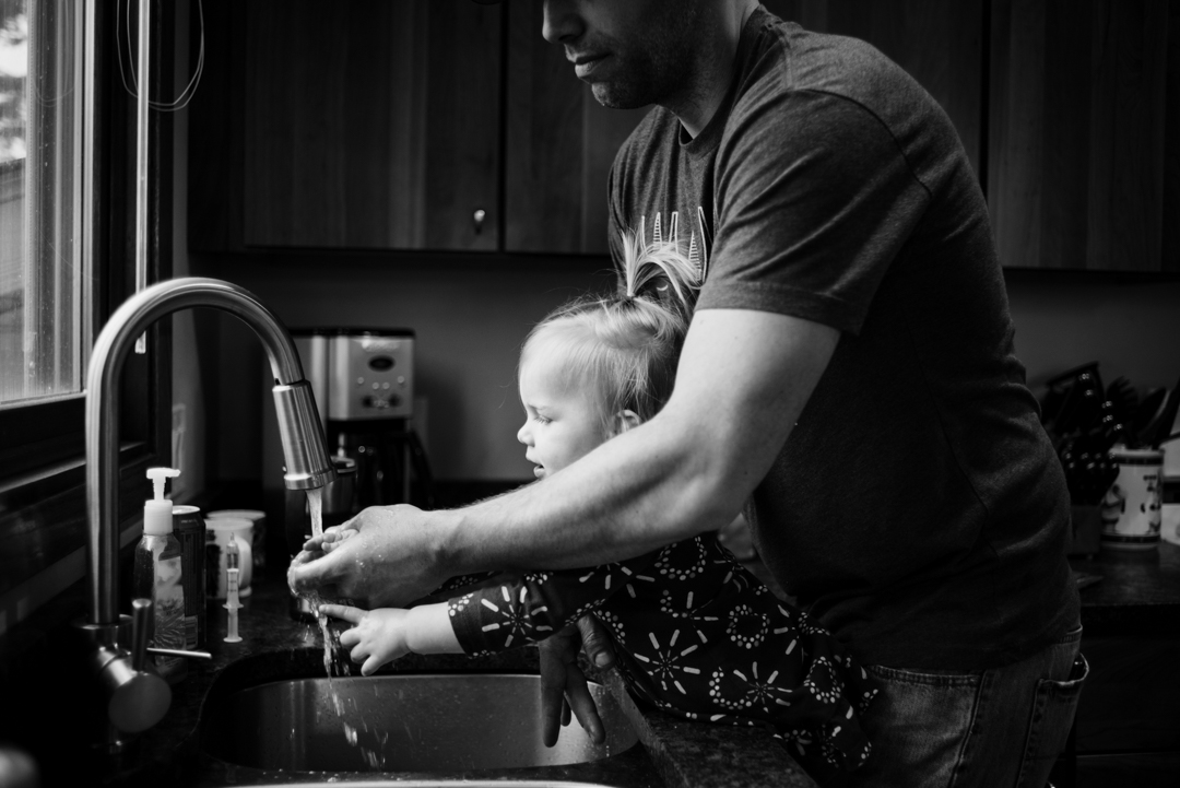 Father helps daughter wash hands at sink.