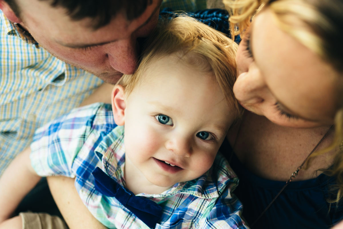 Young boy looks up to camera while mom & dad kiss his cheeks.