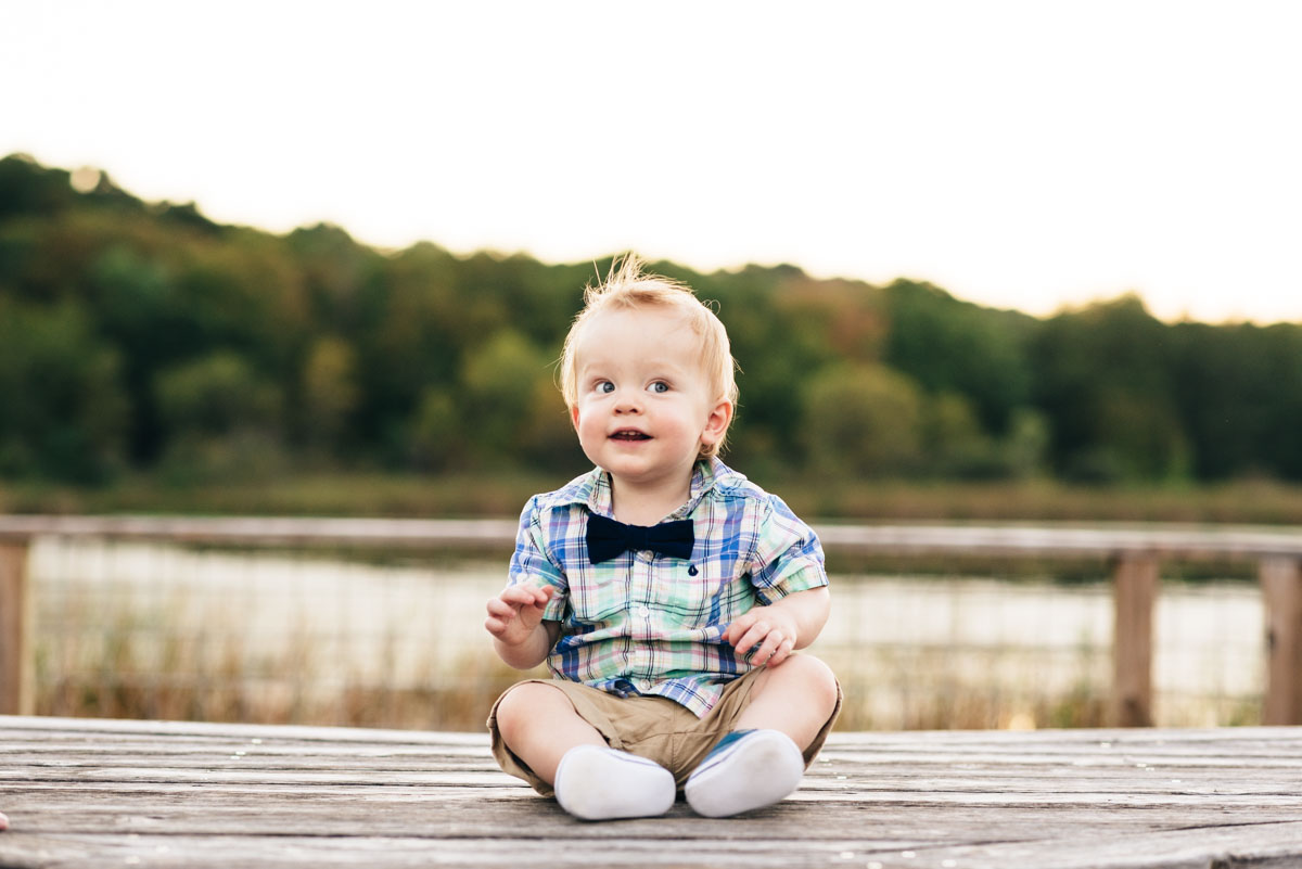 Young boy smiles and sits on bench with lake behind him.