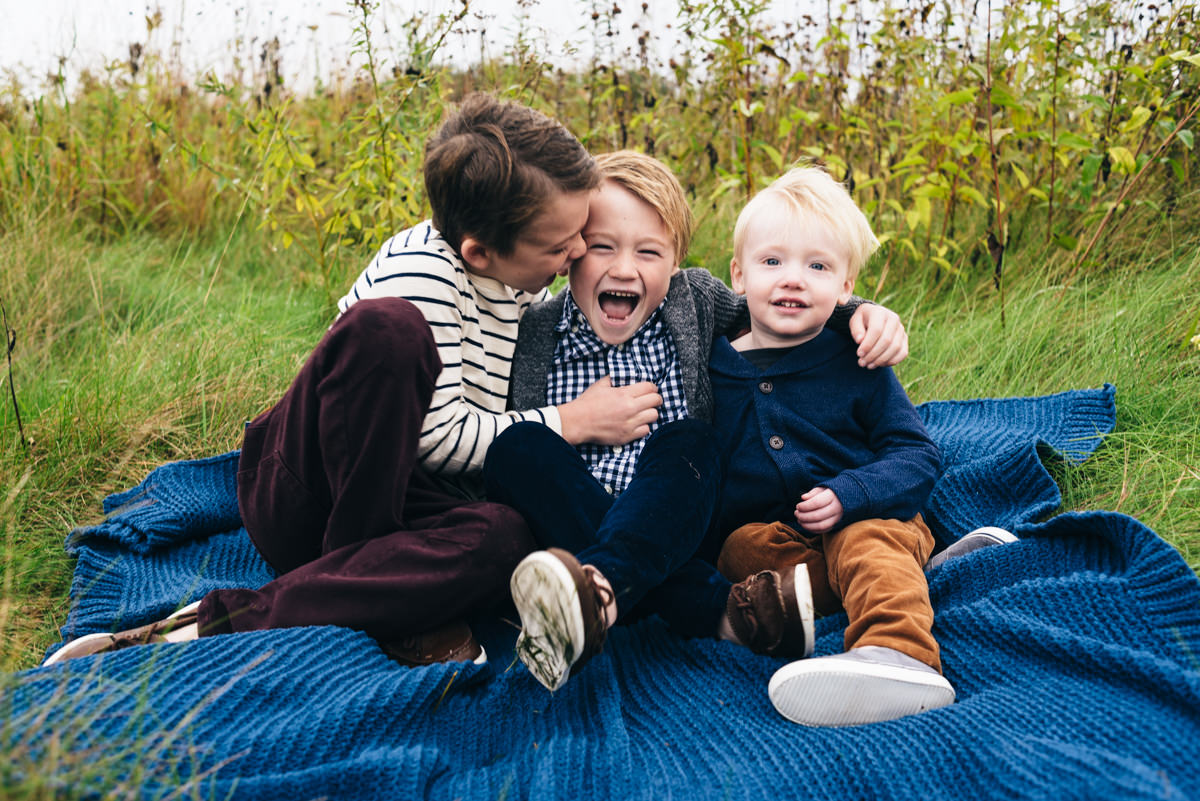 Three young brothers sit on a blanket and tickle each other.