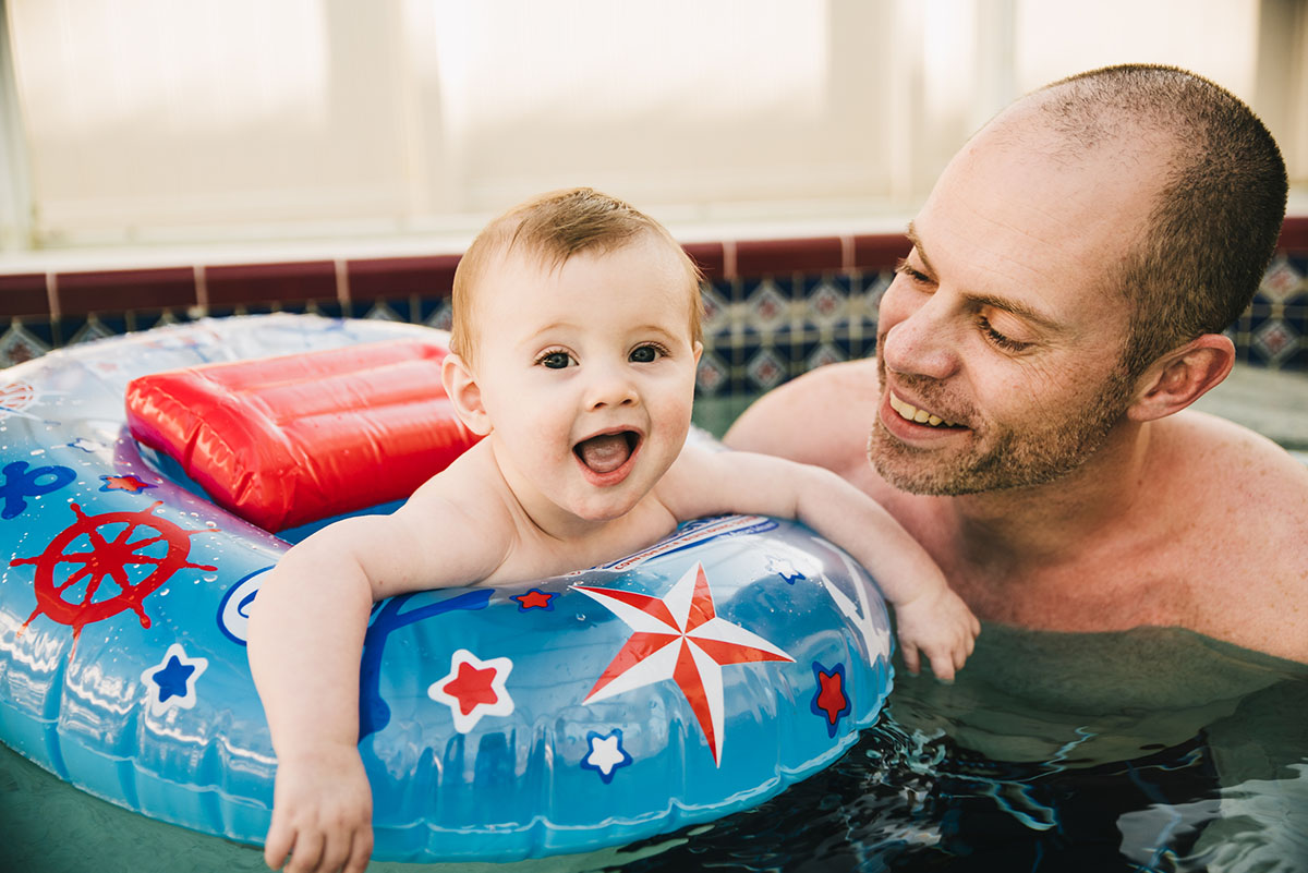 Little boy smiles in pool float while dad looks on. Traveling family photographer.