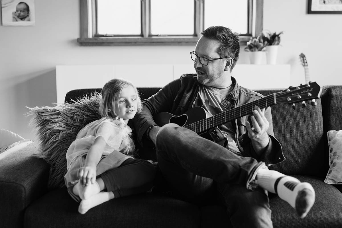 Father and daughter play guitar and sing on a couch.