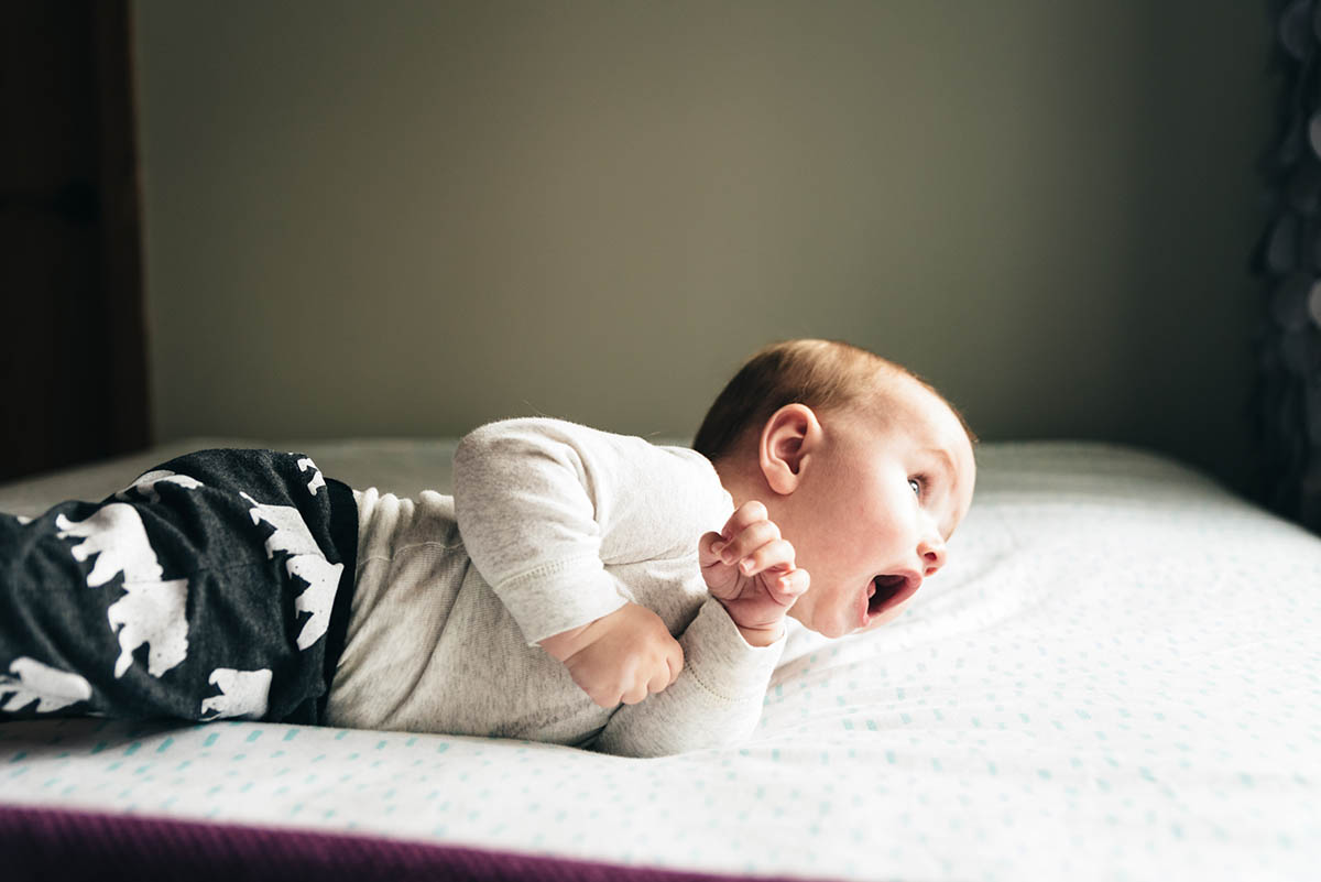 Baby boy looks out a window laying on stomach.