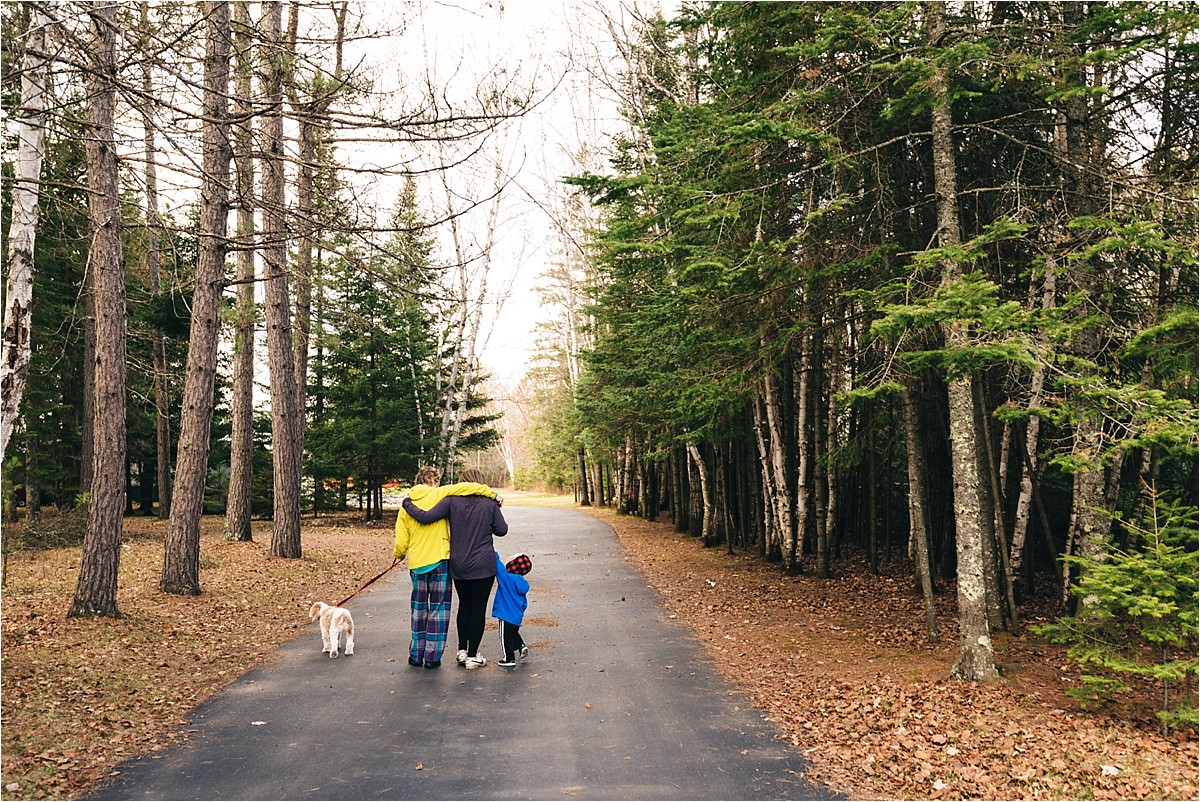 Mother and daughter walk arm in arm down driveway surrounded by pine trees in father's day tribute photo.