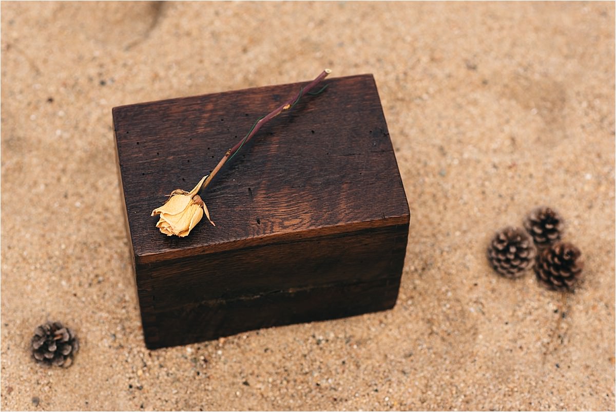 Box of ashes sits on beach with rose on top.