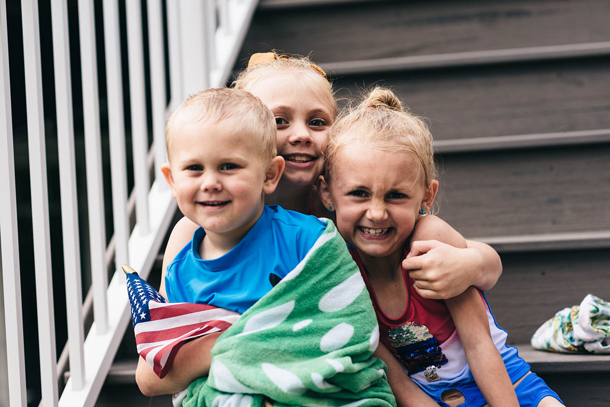 Three kids sit on steps wrapped in towels holding American flags smiling.