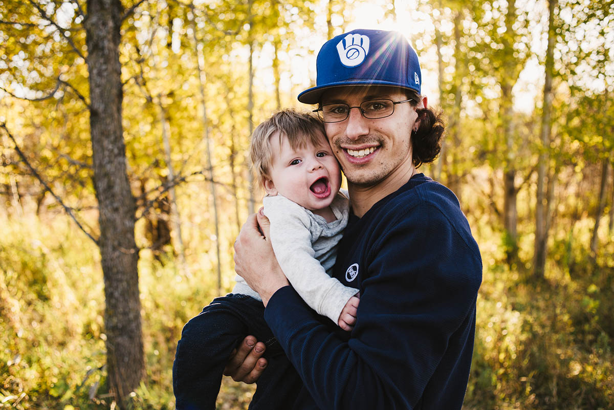 Father snuggles with son as they both smile at the camera in the woods.