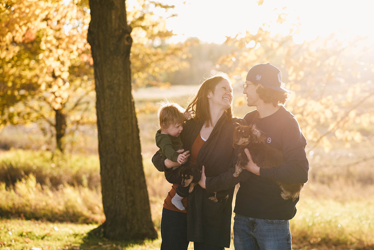 Couple and toddler smile with sunset behind them in woods. Sunset family session.