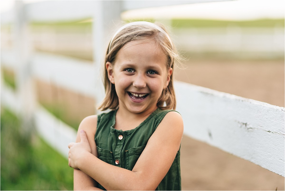 Young girl in green dress laughs at the camera near a white fence.