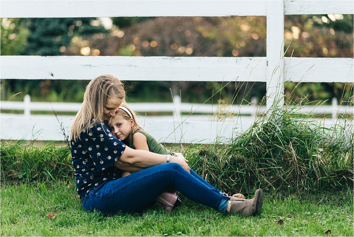 Mother and daughter photoshoot sitting by white fence in tall grass.