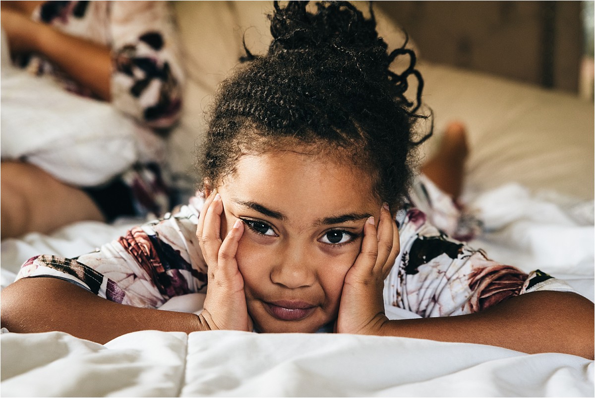 Little girl smiles while she lays on a bed during lifestyle in-home newborn photography session.