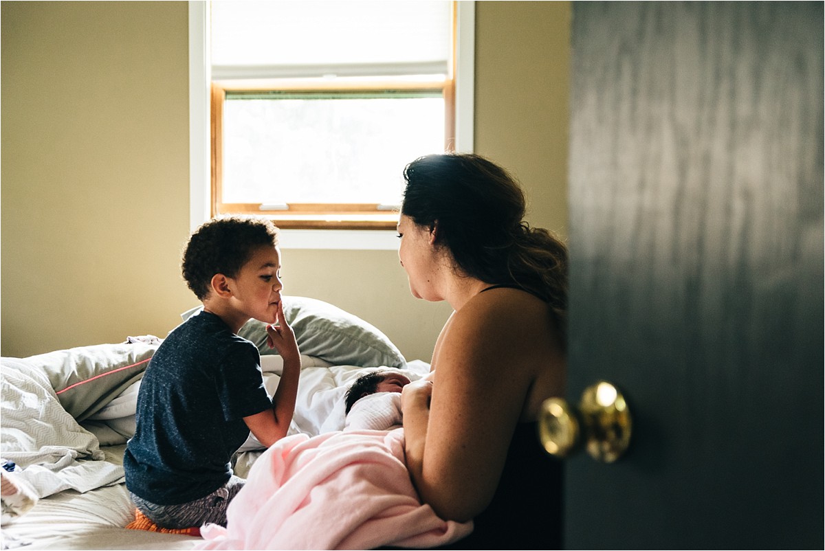 Young boy curiously looks on as mother cradles newborn baby sister.