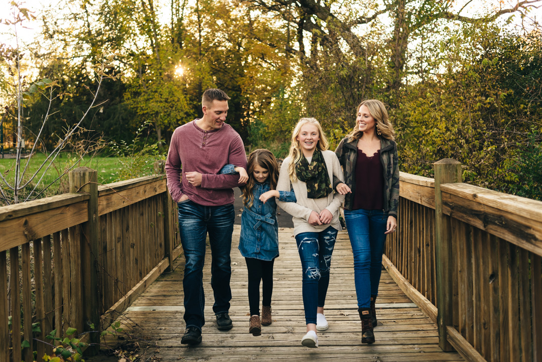 Family of four links arms and walks across bridge during family photos at sunset