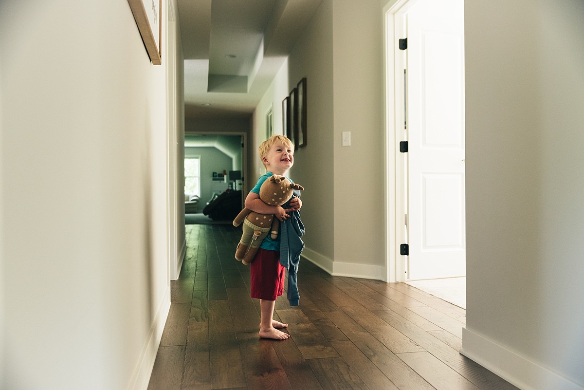 Little boy stands in hallway holding stuffed animal.
