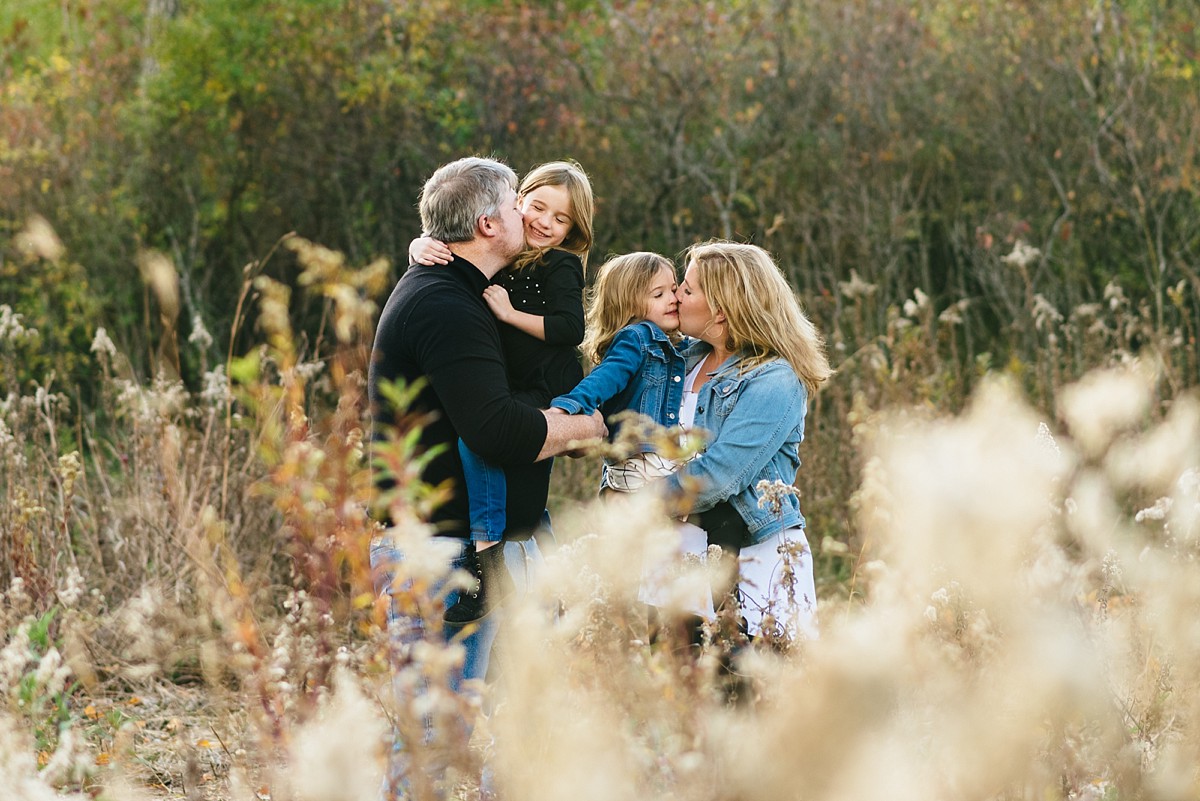 Family of four gives kisses in a field during a sunset photography session.