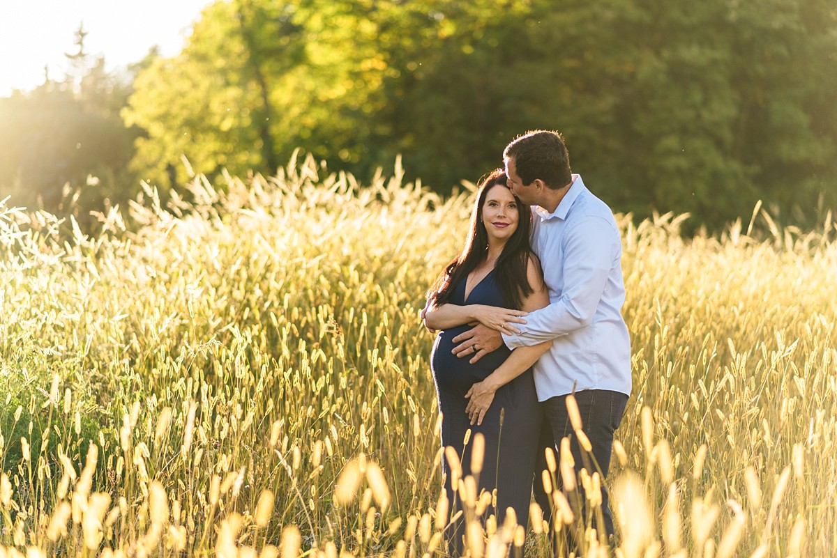 Couple at sunset stands in glowing tall grass. Woman is pregnant and father kisses mother on cheek.