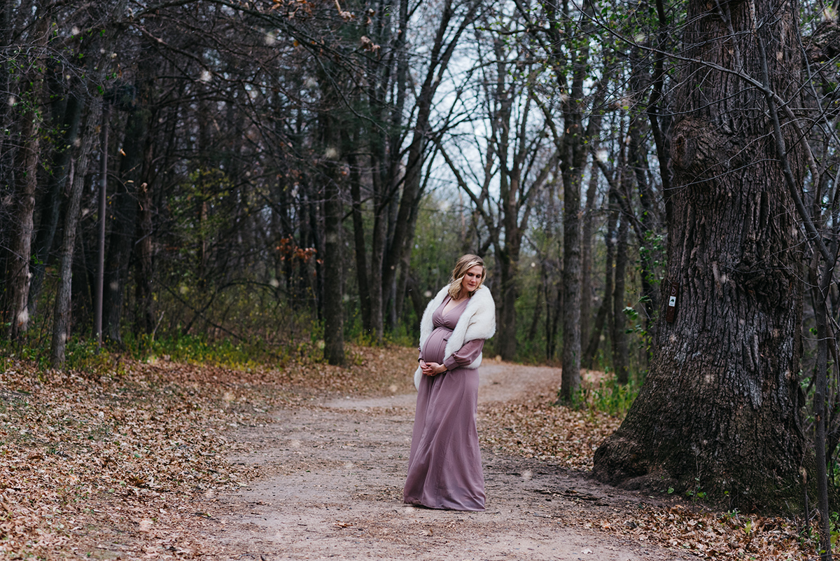 Pregnant woman in beautiful pink dress stands in woods as snow falls.