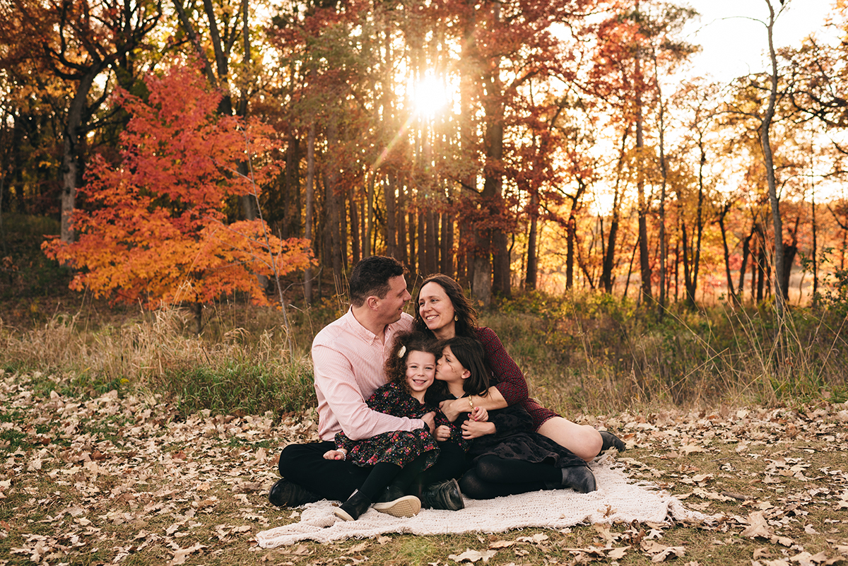 Family of four sit on blanket in the sunset laughing with each other.