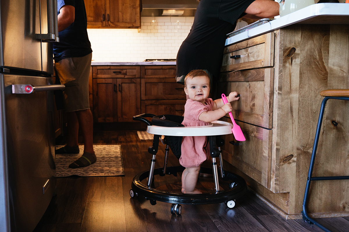 Toddler in wheeler scoots around kitchen while her family cooks.