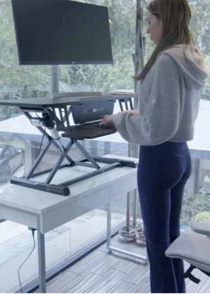 A woman working at a standing desk with a monitor and a keyboard in a bright, modern home office setting. A woman working at a standing desk with a monitor and a keyboard in a bright, modern home office setting.