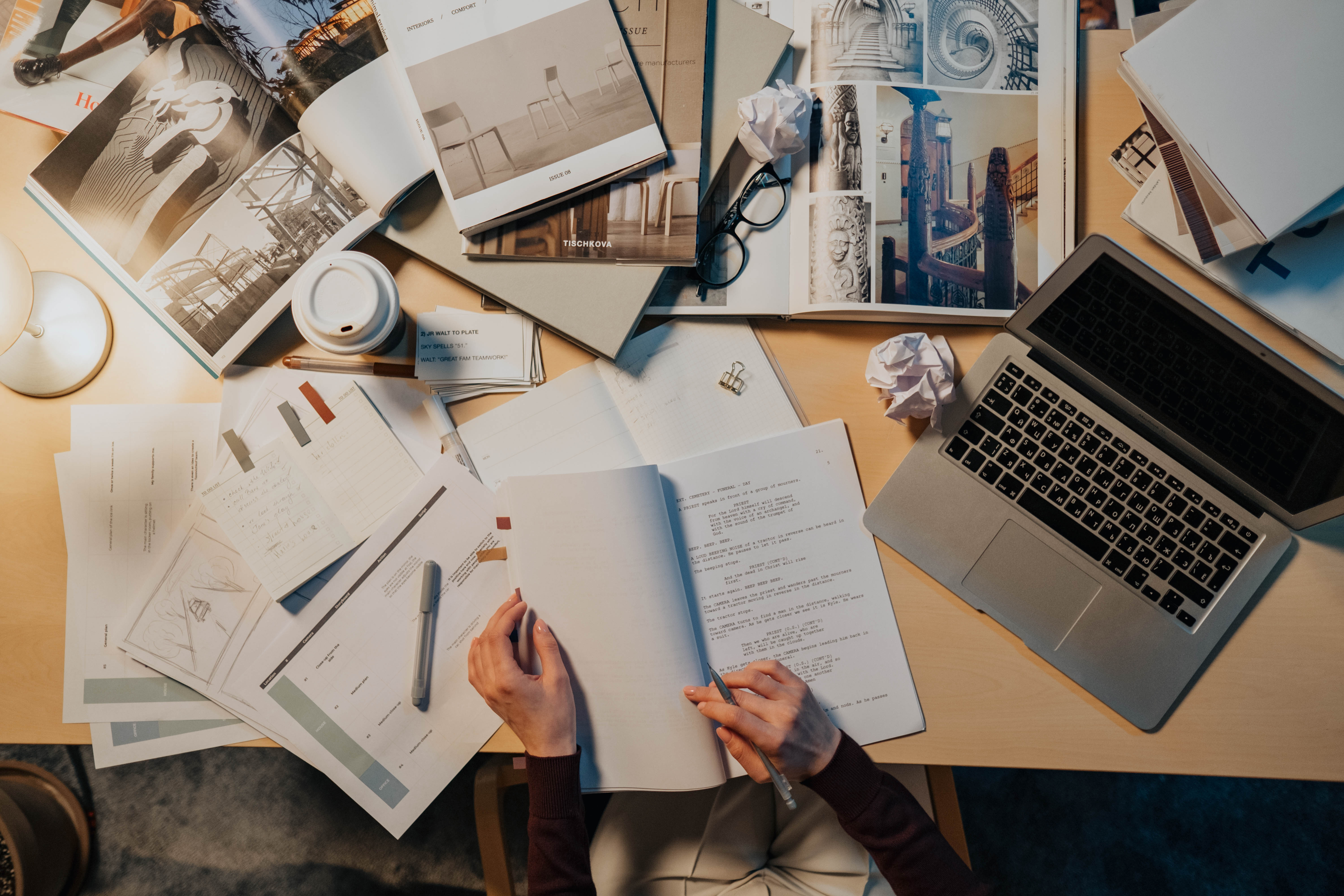 A cluttered wooden desk filled with open books, papers, a laptop, and a coffee cup. A cluttered wooden desk filled with open books, papers, a laptop, and a coffee cup.