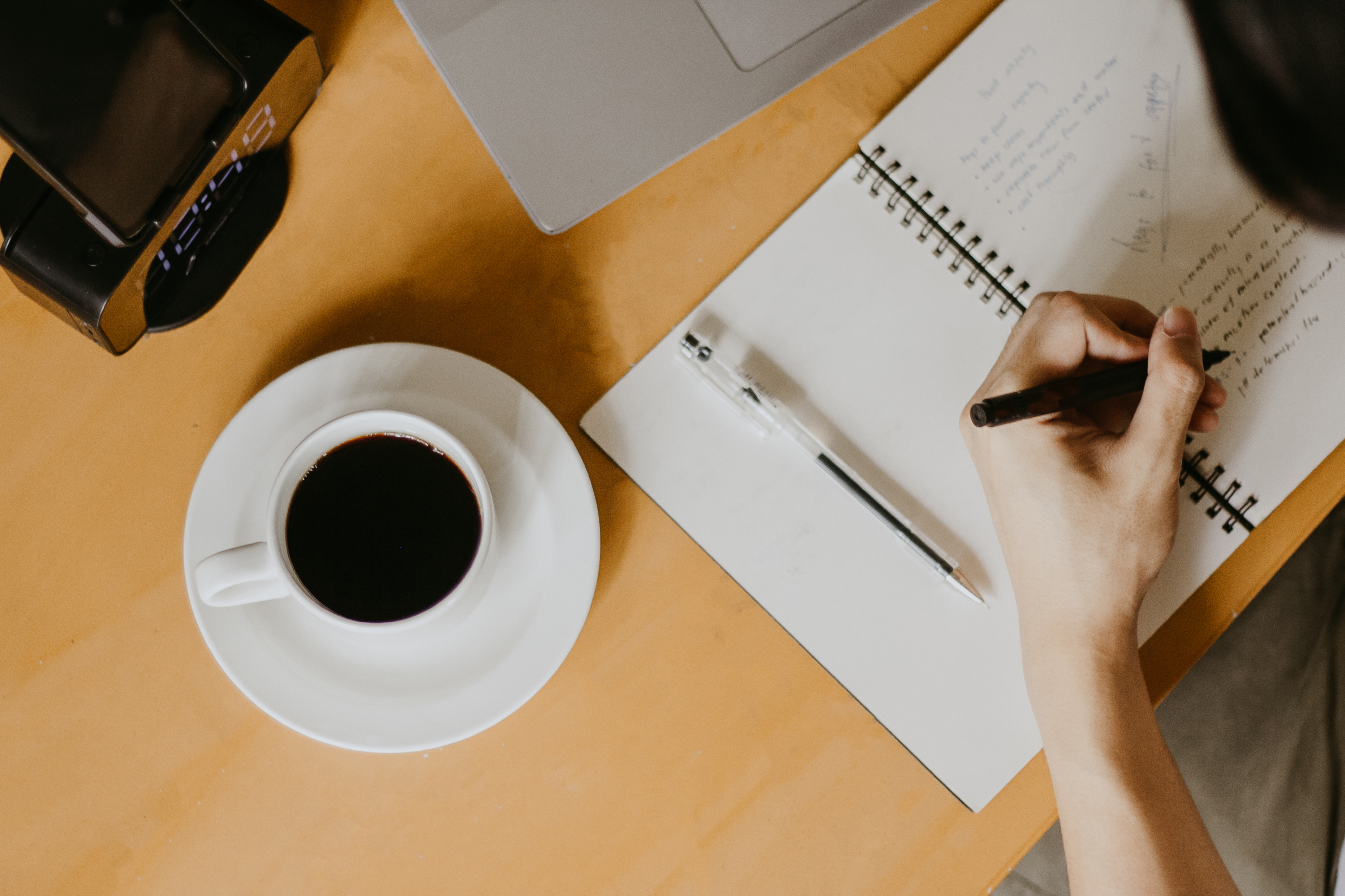 Person writing in a spiral notebook beside a cup of coffee and laptop—depicting writing tools and workspace for authors.