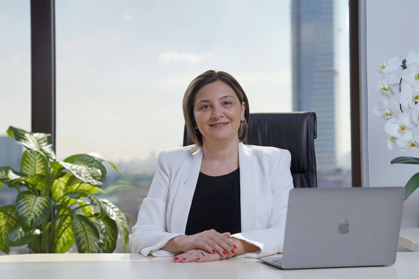 Confident professional woman in white blazer sitting at a modern office desk with laptop and plants, representing successful interview preparation and executive presence