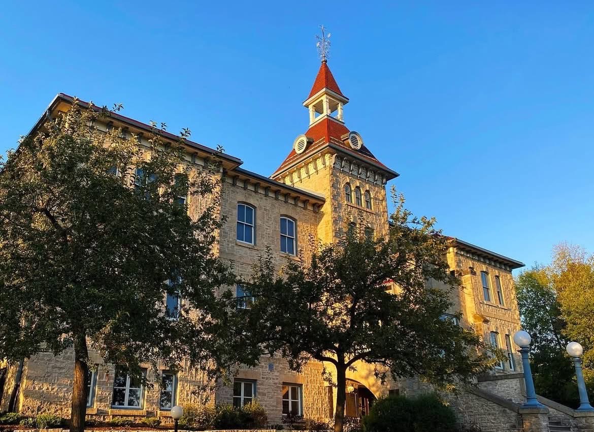 Historic stone building with a red-roofed clock tower and large windows, surrounded by trees under a clear blue sky at sunset. Historic stone building with a red-roofed clock tower and large windows, surrounded by trees under a clear blue sky at sunset.