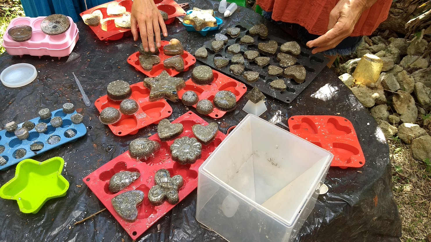 Hands molding various shapes of rocks or concrete in silicone molds, including stars, hearts, and flowers, on a table outdoors. Hands molding various shapes of rocks or concrete in silicone molds, including stars, hearts, and flowers, on a table outdoors.