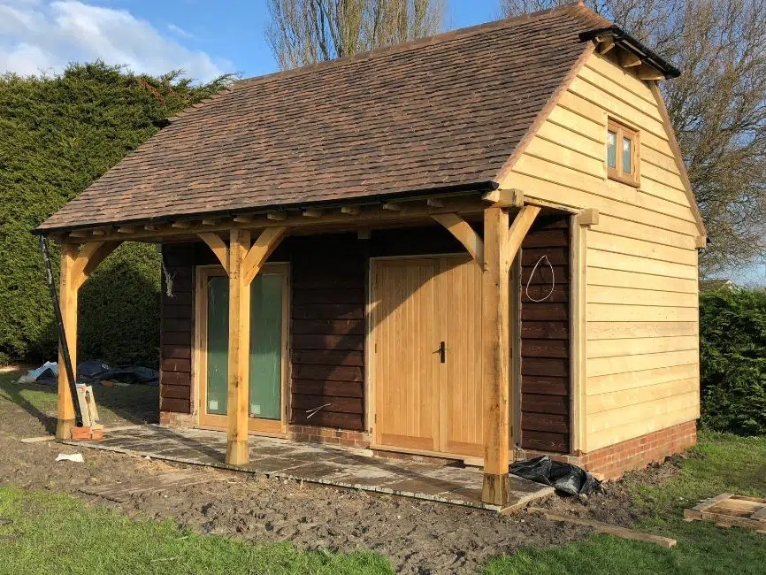 Oak-framed garden room with wooden structure, sloped roof, and front porch, surrounded by greenery, illustrating outdoor living space options.