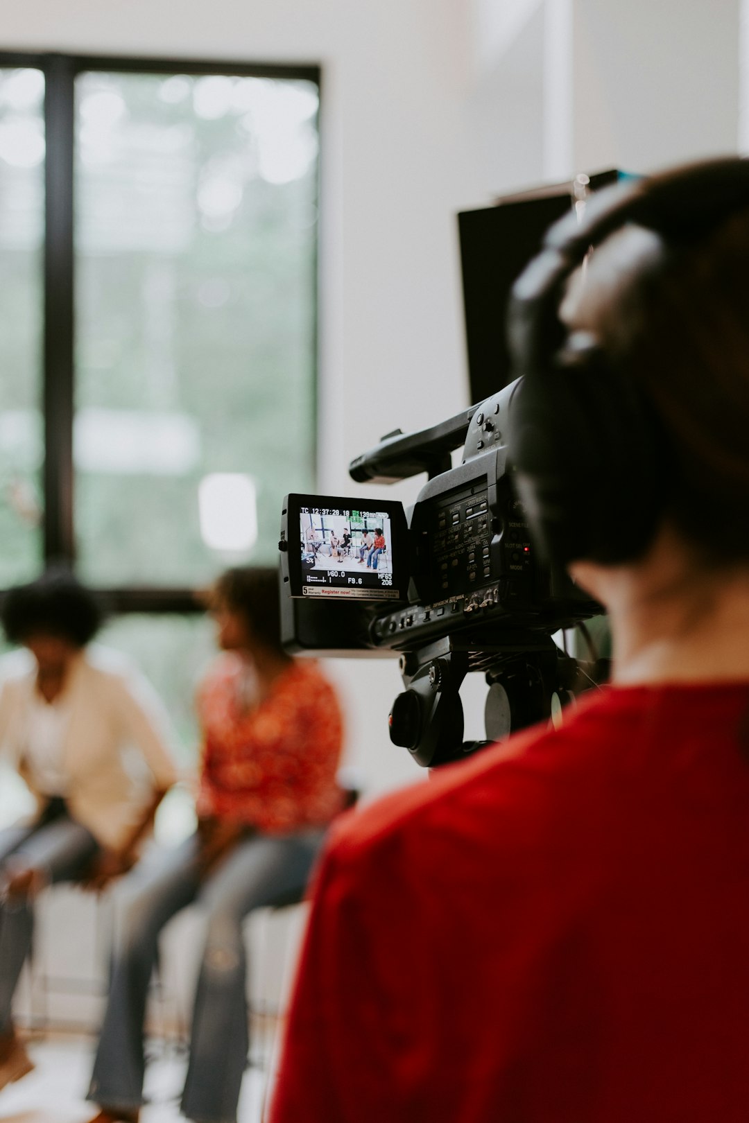 Camera operator filming two women seated indoors for an interview or discussion, with the subjects visible on the camera's viewfinder screen. Camera operator filming two women seated indoors for an interview or discussion, with the subjects visible on the camera's viewfinder screen.