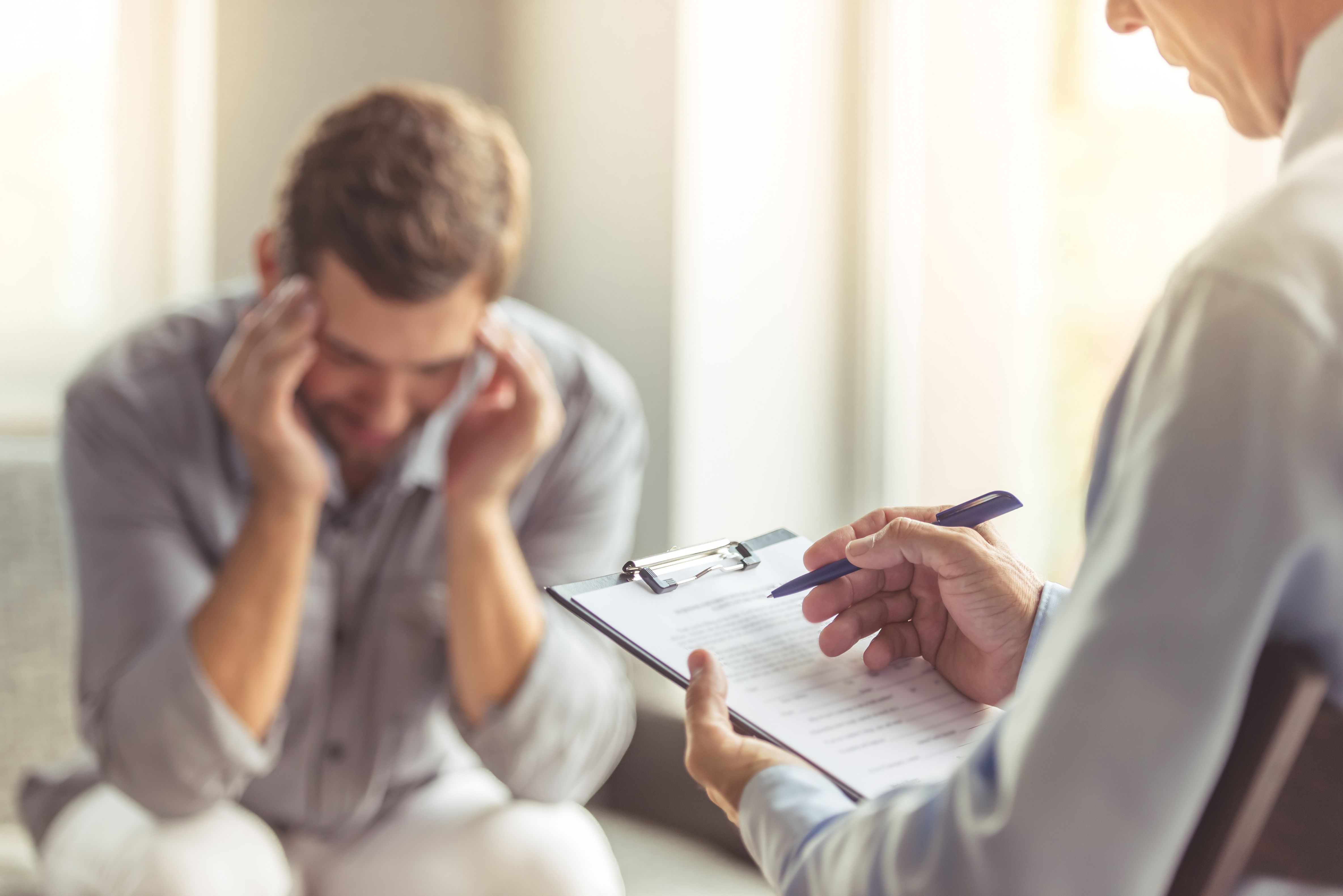 Depressed man in casual clothes is leaning on his knees and telling about his problems while sitting on the couch at the psychotherapist.