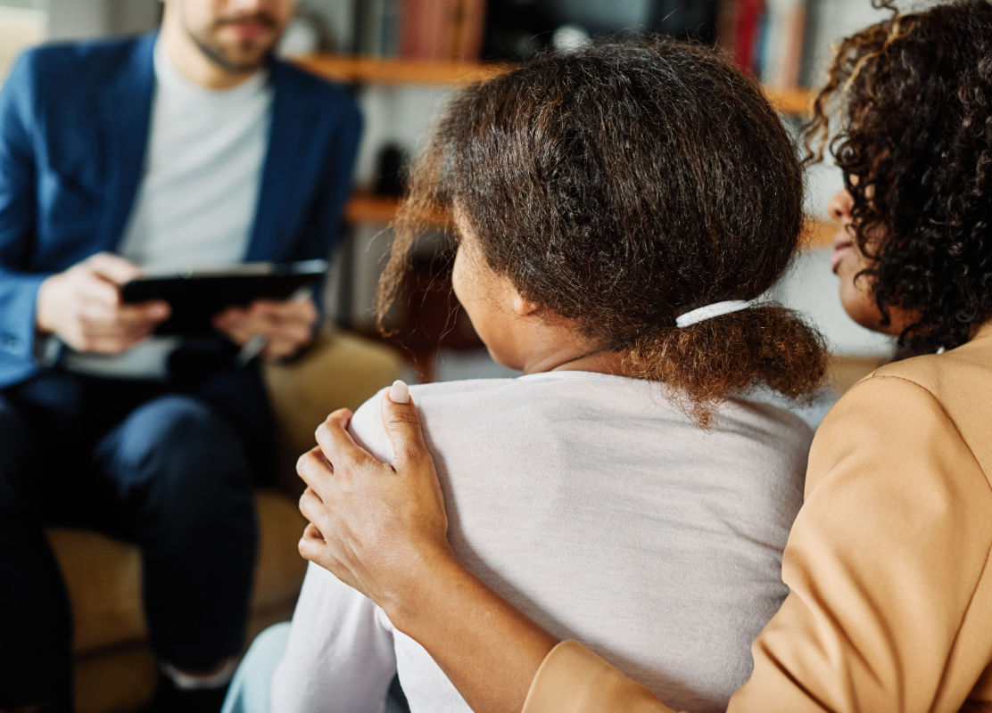 Woman wraps arm around a young girl, in the background a man sits in a chair with a clipboard