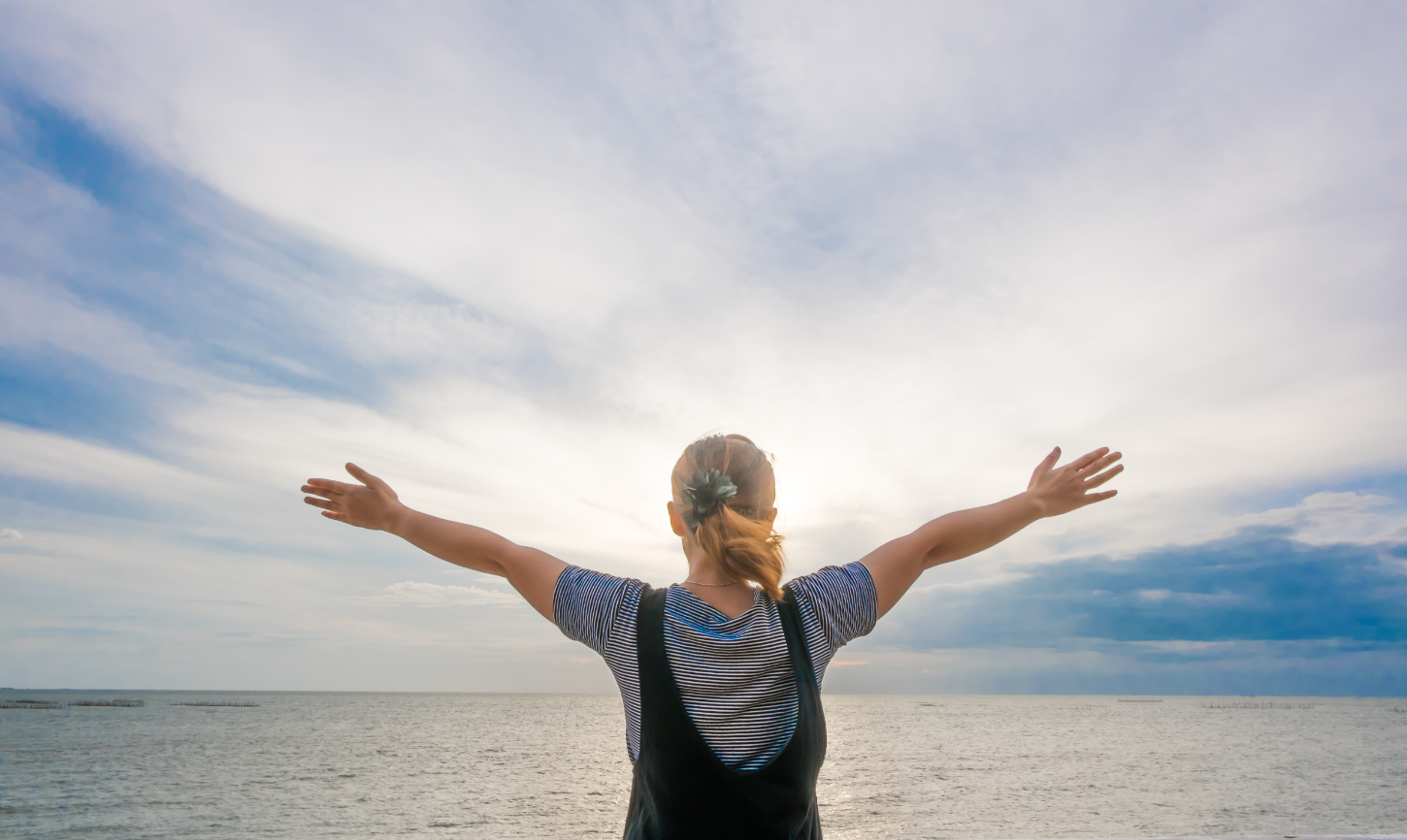 Image of the back of a woman wearing overalls with arms outstretched towards the sky